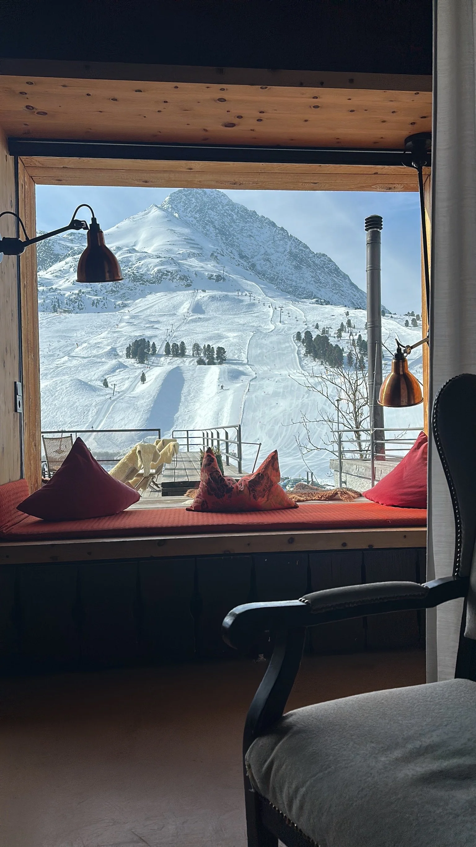 Snow-covered mountain view from a room with a window seat decorated with pillows and blankets, overlooking ski slopes and chairlifts.