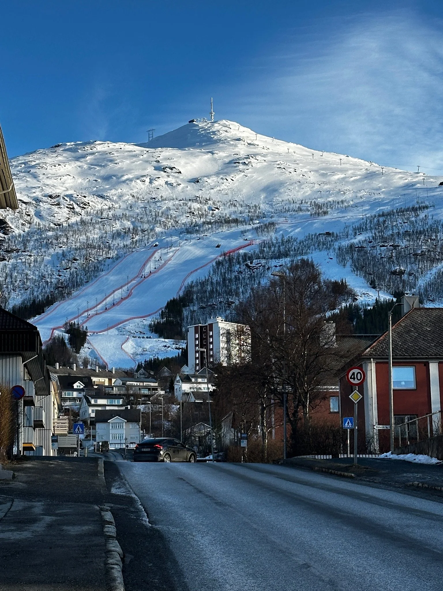 A residential neighborhood with a snow-covered mountain in the background, showing ski slopes and ski lift poles. The street is wet with some patches of snow, and houses with cars parked outside are visible. A speed limit sign indicates 40 km/h.