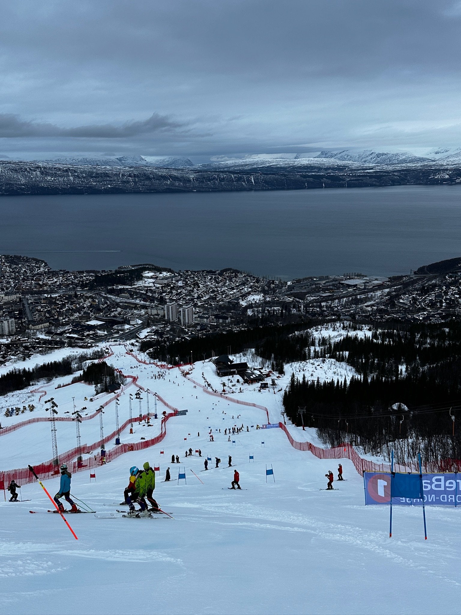 People skiing down a snowy slope at a ski resort, with a town and snow-covered mountains in the background under a cloudy sky.