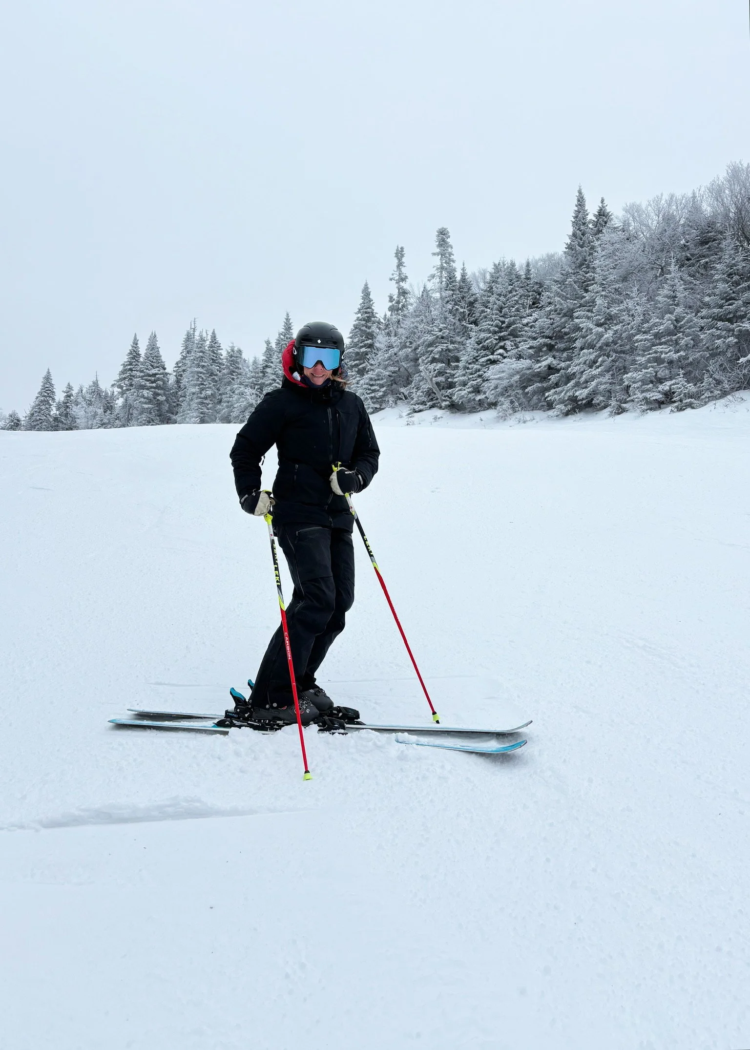 Woman in black ski gear, helmet, and goggles skiing on snow-covered landscape with pine trees in the background.