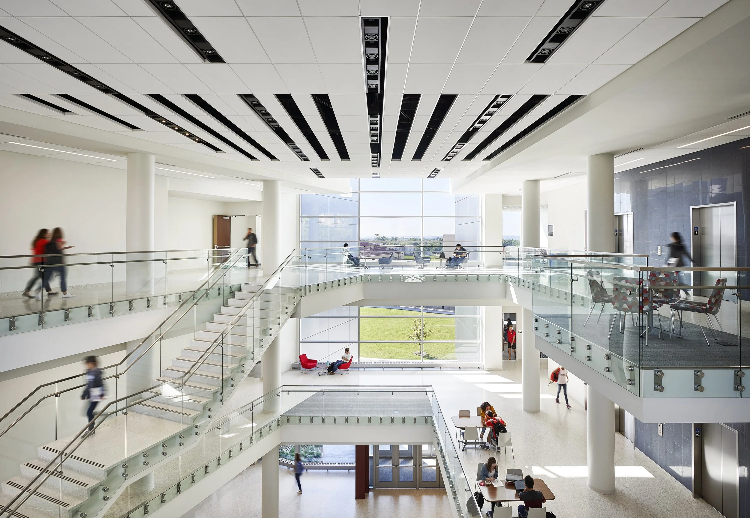 Modern multi-level inside building with people walking, sitting, and using elevators, glass railings, large windows, and indoor staircases.