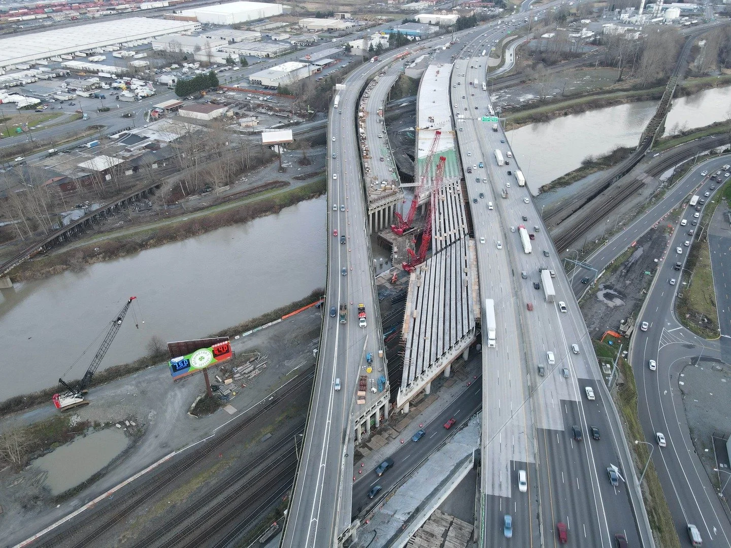 Aerial view of a highway construction site with cranes, partially built bridges, and traffic, near a river with industrial and commercial areas in the background.