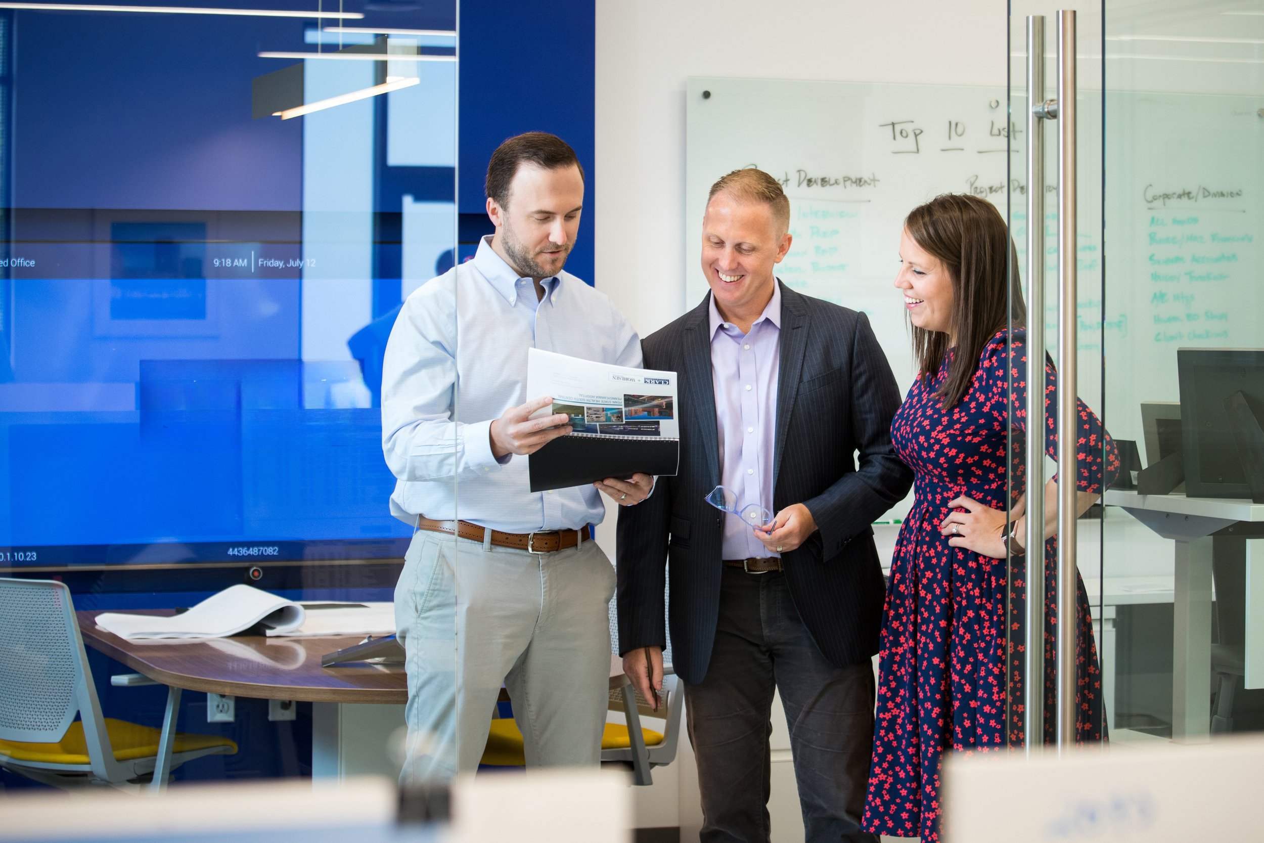 Three business professionals, two men and one woman, standing together in an office, looking at a document and smiling.