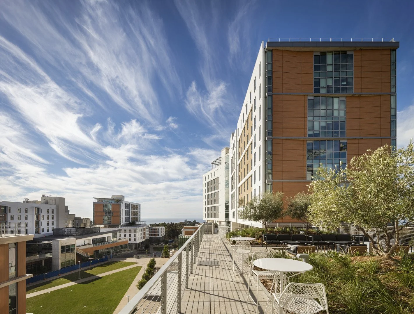 View from a rooftop terrace in an urban area, showing modern high-rise buildings, a green landscaped area, and a blue sky with wispy clouds.