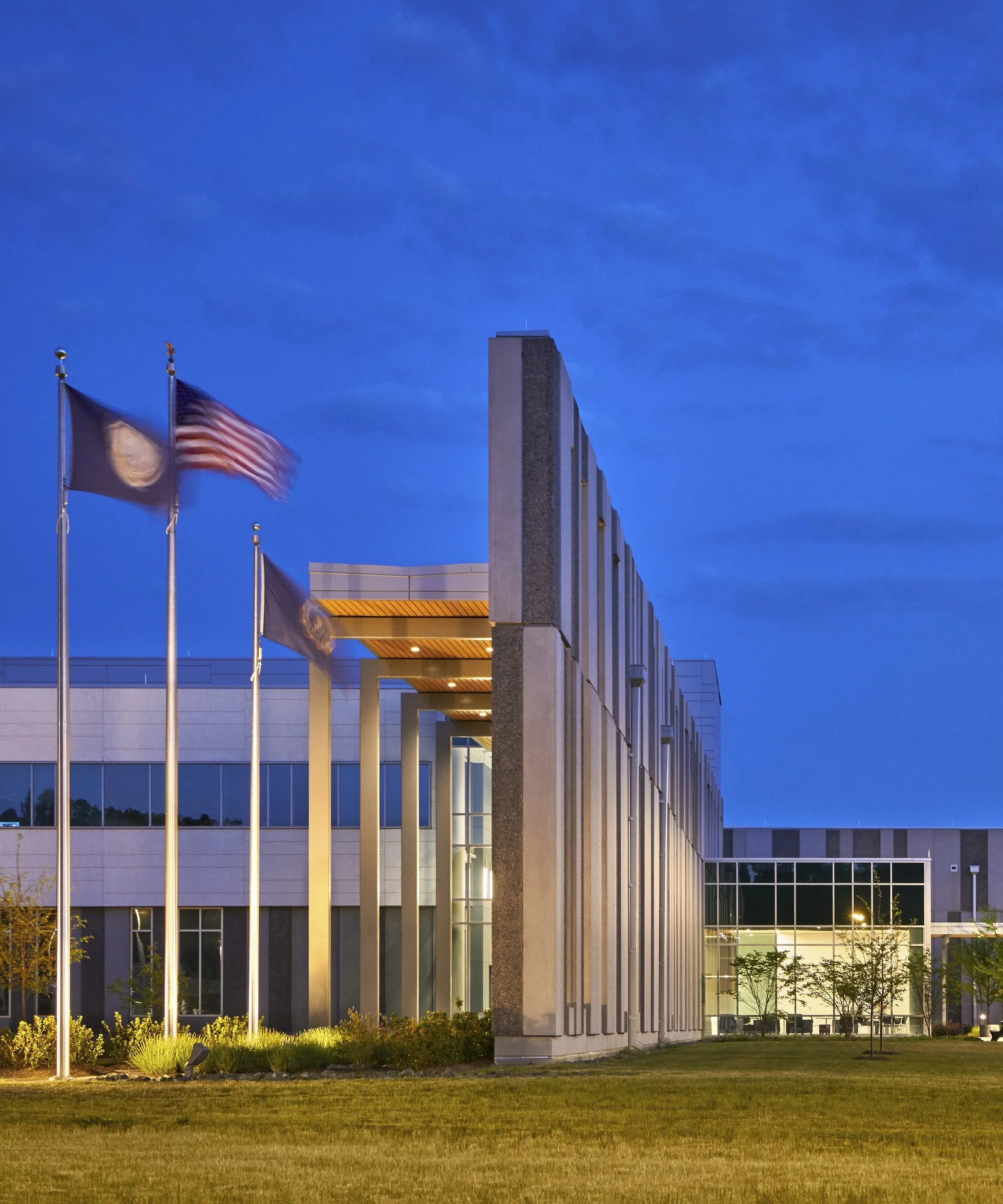 Modern building with flags on poles, illuminated at sunset or dusk, with a grassy area and small trees in front.
