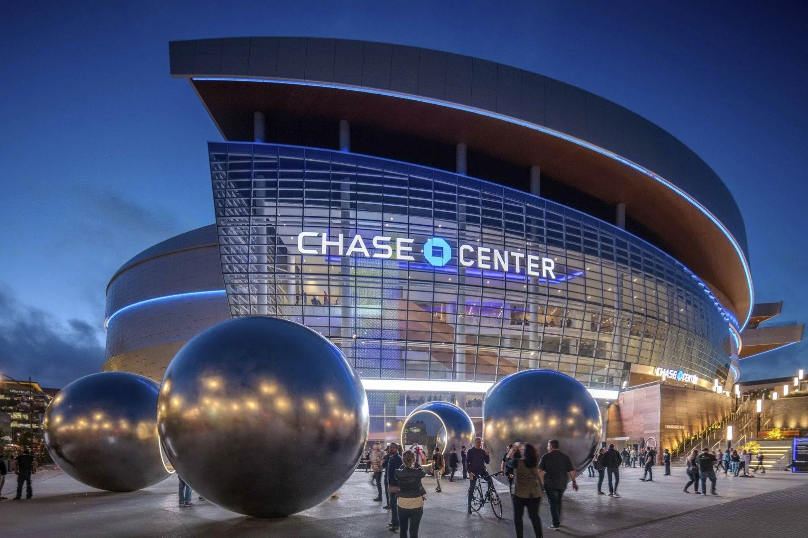 Night view of the Chase Center, a modern sports arena, with illuminated signs and large metallic spheres outside, and people walking around.