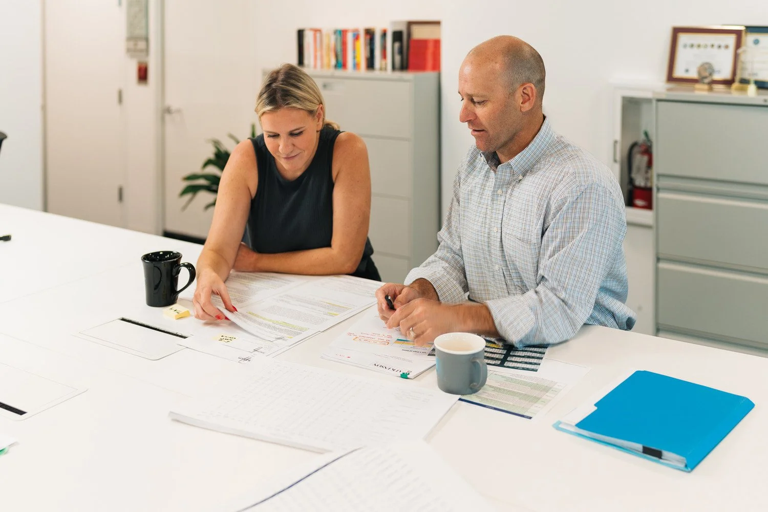Two people, a woman and a man, sitting at a white table in an office, reviewing documents and engaging in a discussion.