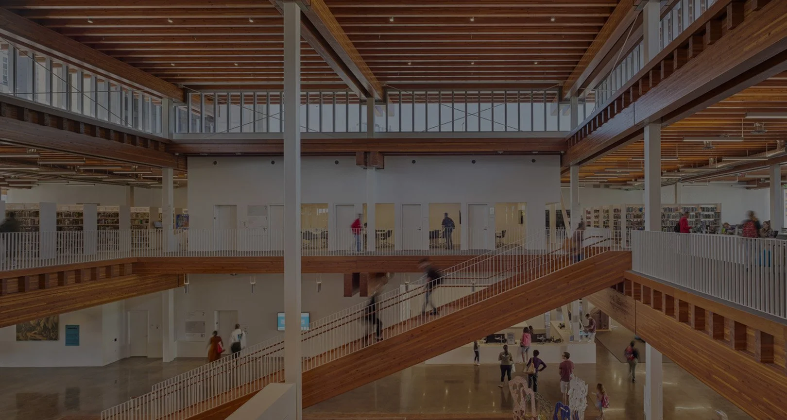 Interior view of a modern multi-level library with wooden railings and escalators, people walking and browsing shelves.