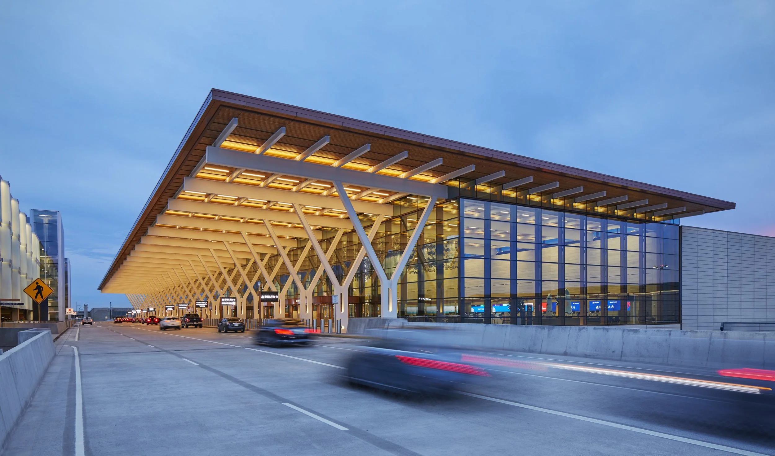 Modern airport terminal with glass walls and wooden roof, cars driving on the road, and a twilight sky.