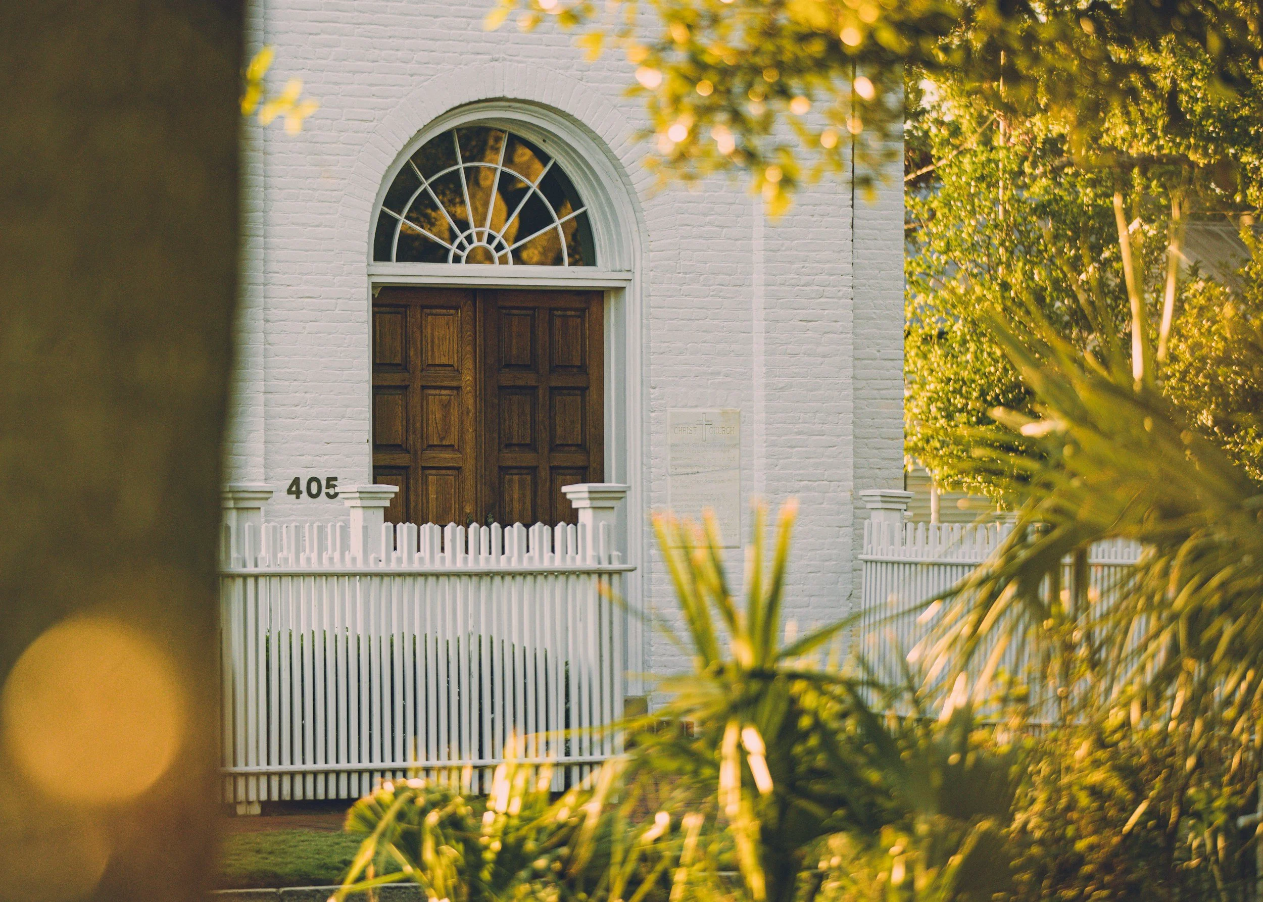 A white brick church with a wooden door, a semi-circular window above the door, a white picket fence in front, the house number 405 displayed, and trees with green leaves surrounding it, captured during golden hour.