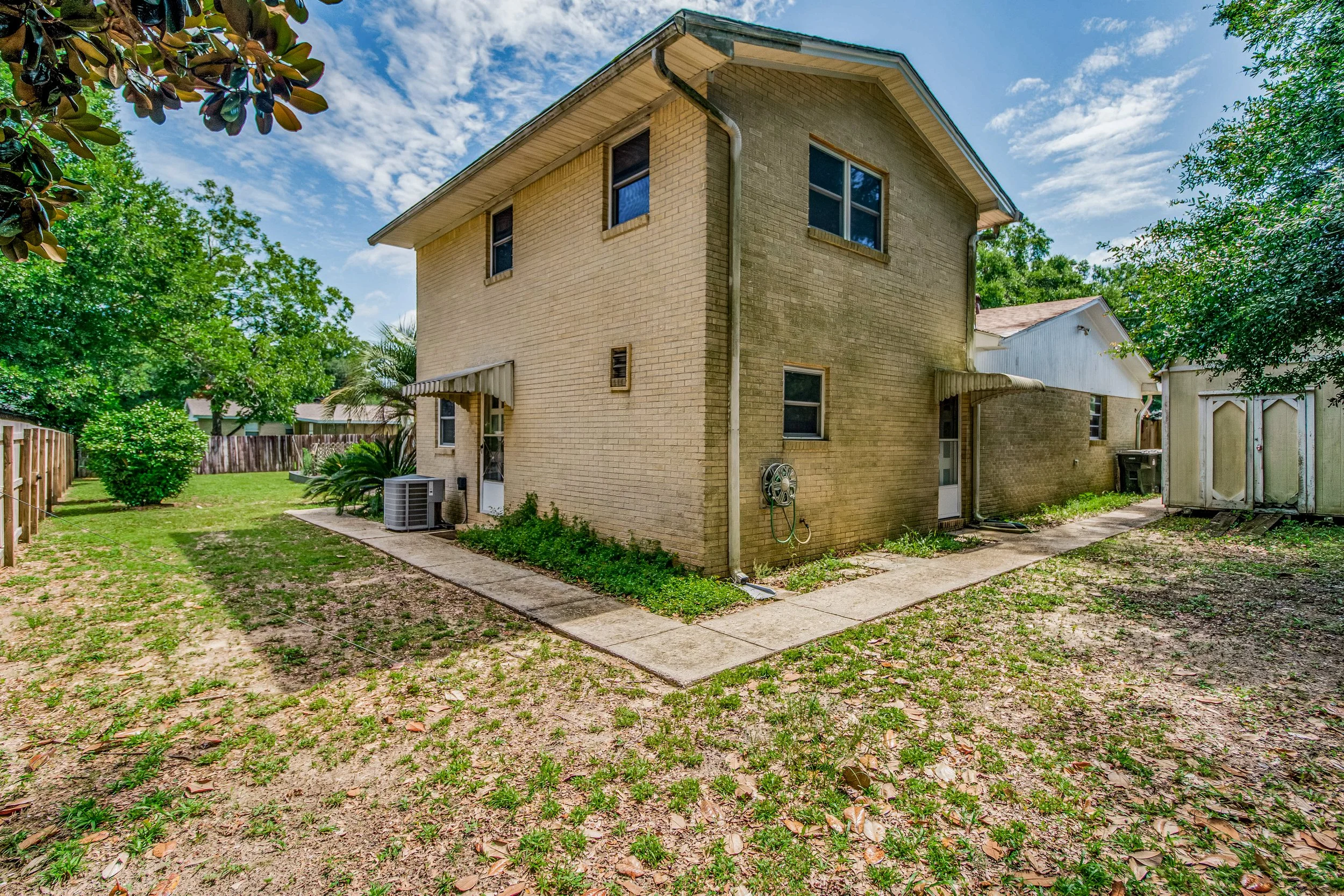 Backyard view of a two-story beige brick house with a small concrete pathway and grassy area, surrounded by trees and a wooden fence.