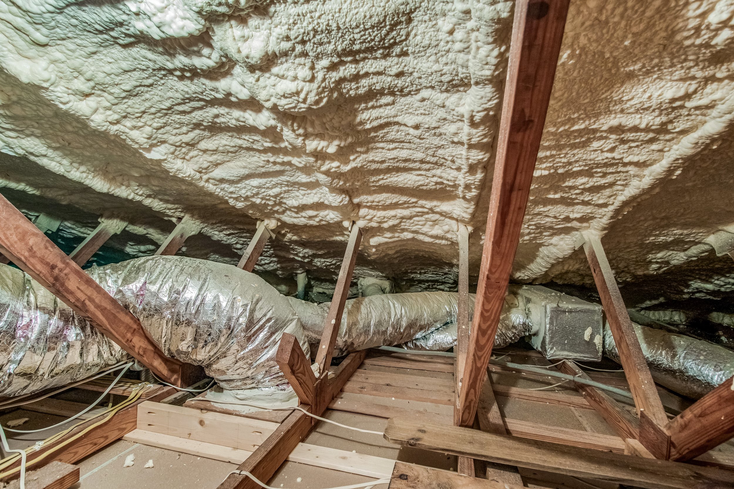 Insulation foam spray on the ceiling of an attic space with visible wooden beams and HVAC ductwork.