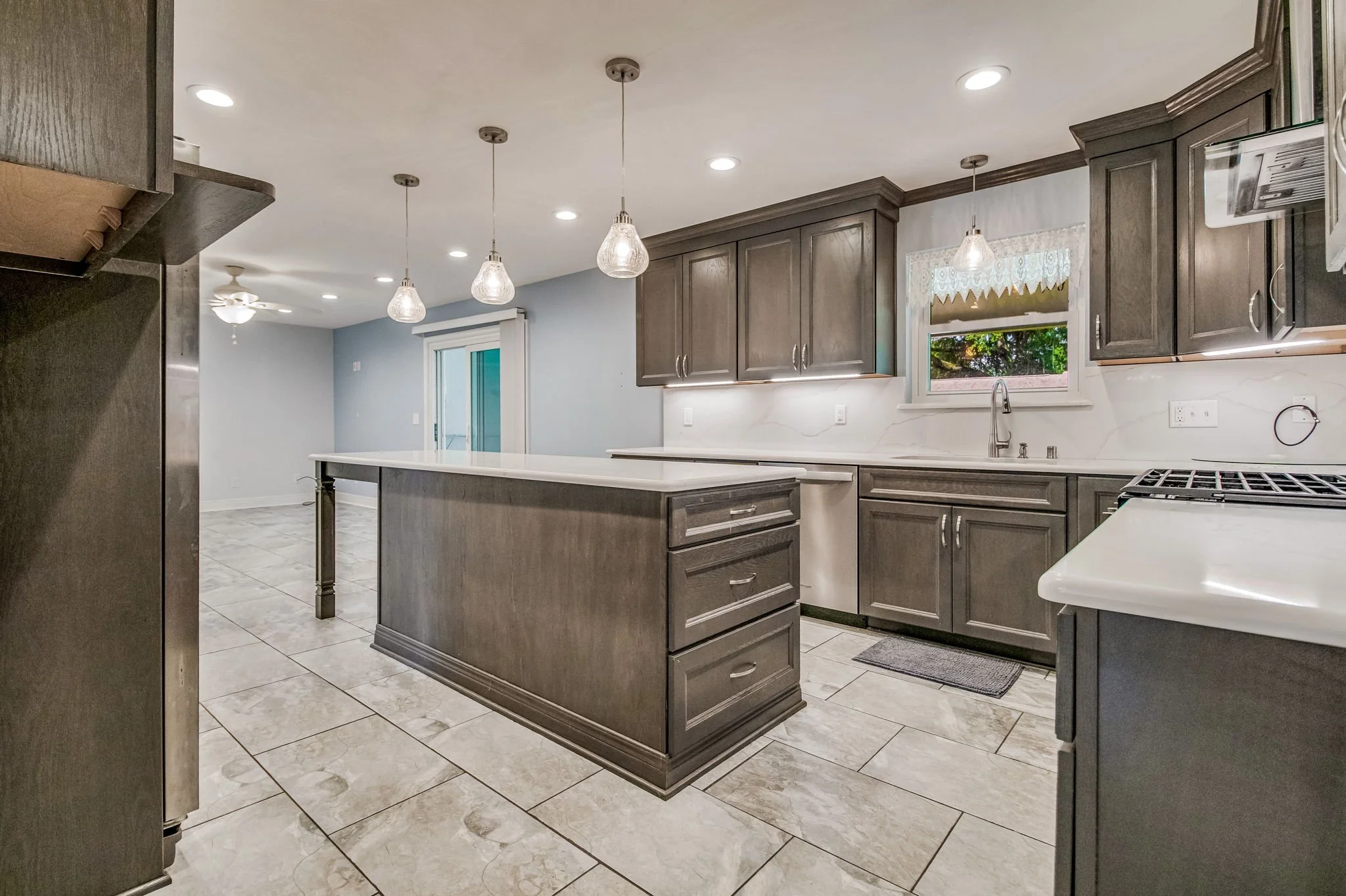 Modern kitchen with gray cabinets, white countertops, pendant lights, tiled floor, window above sink, slide door in background.