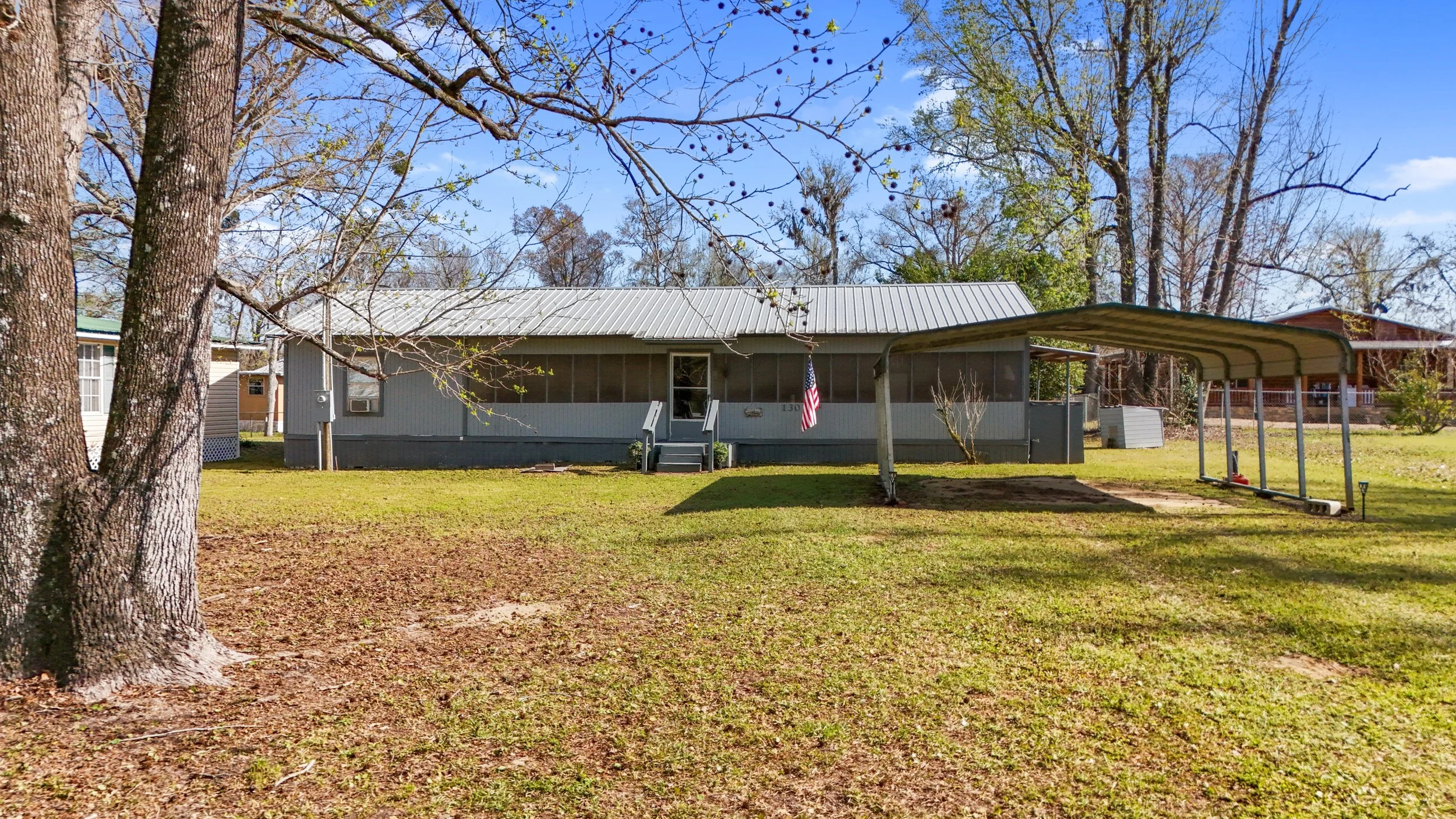 A single-story gray house with a metal roof, a front step, and an American flag displayed outside. The yard has a tree with few leaves and a carport on the right. The sky is blue with scattered clouds.