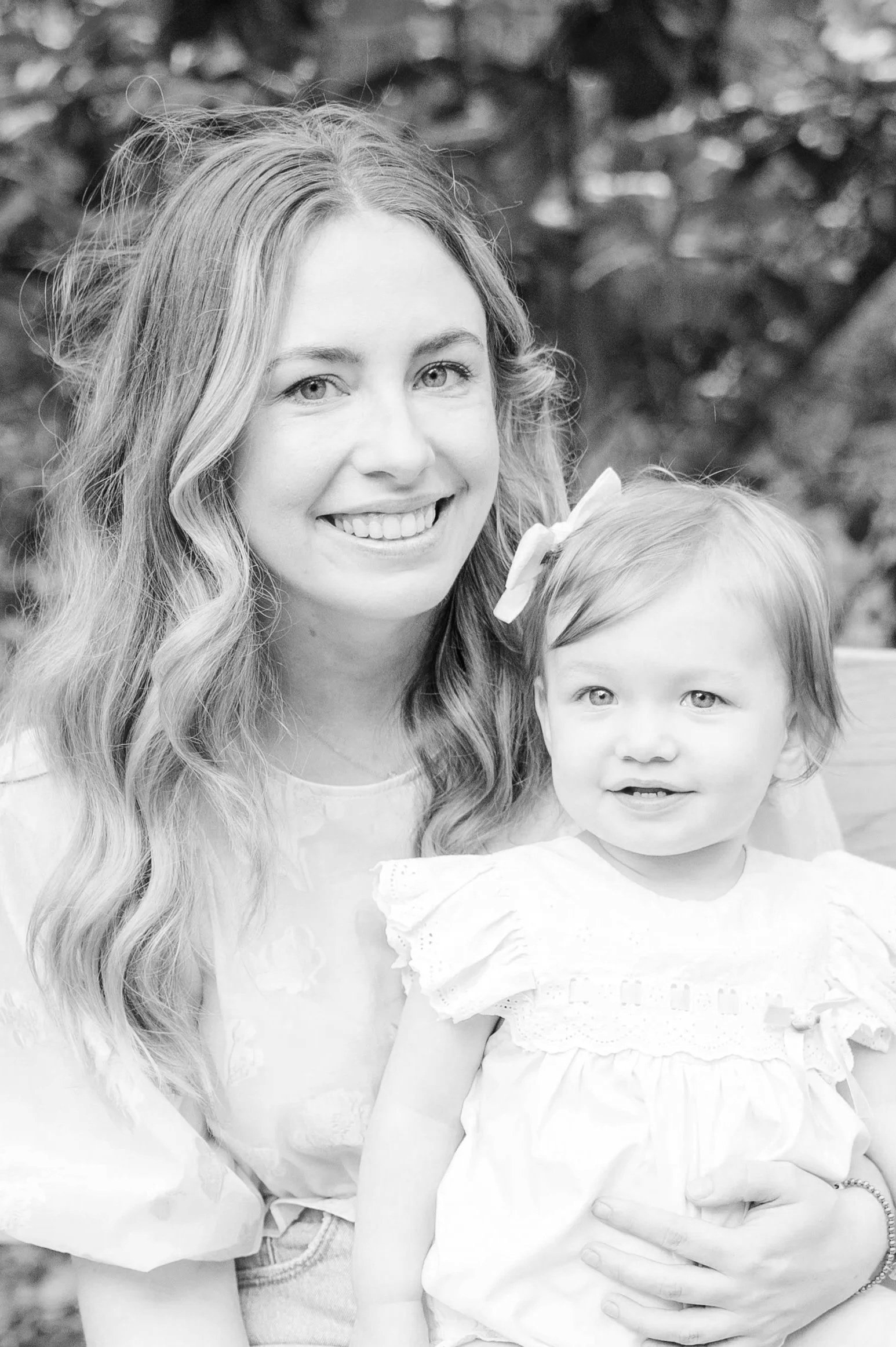 A woman with long wavy hair smiling while holding a young girl with a bow in her hair, both outdoors against a blurred natural background.