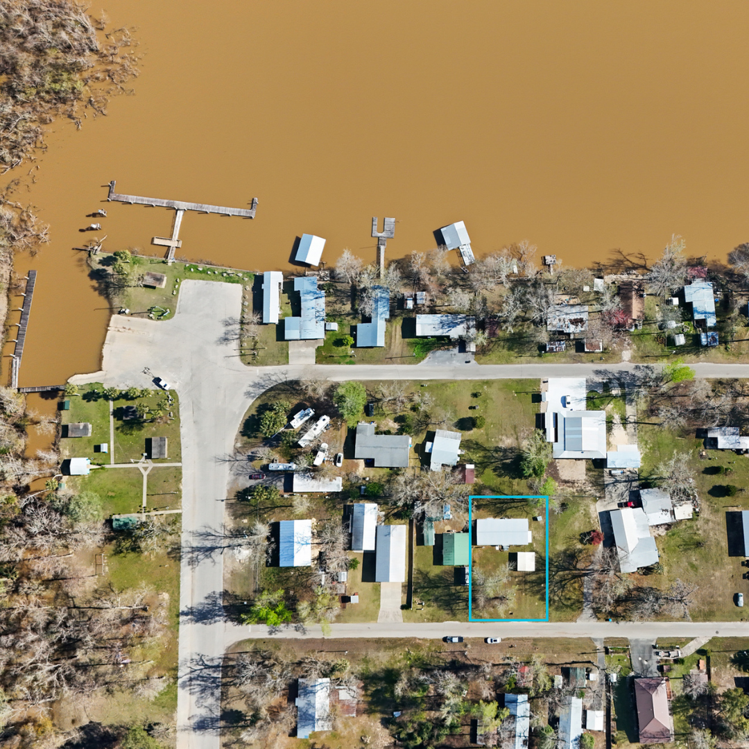 Aerial view of a neighborhood near a brown river, showing houses, trees, and roads, with a highlighted property outlined in blue.