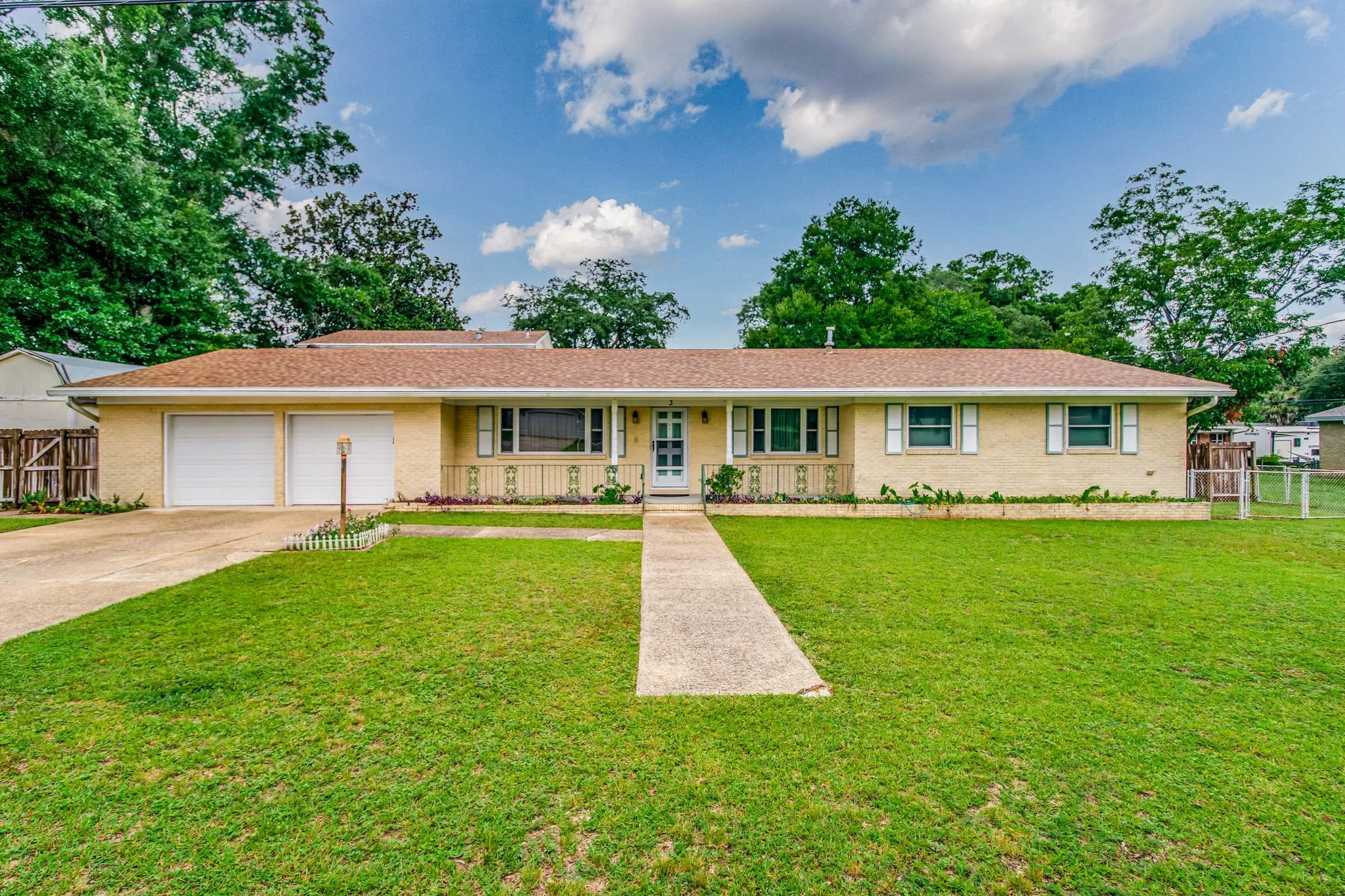 Front view of a single-story, yellow brick house with a brown roof, a concrete walkway leading to the front door, a two-car garage on the left, and a green lawn with some small garden beds.