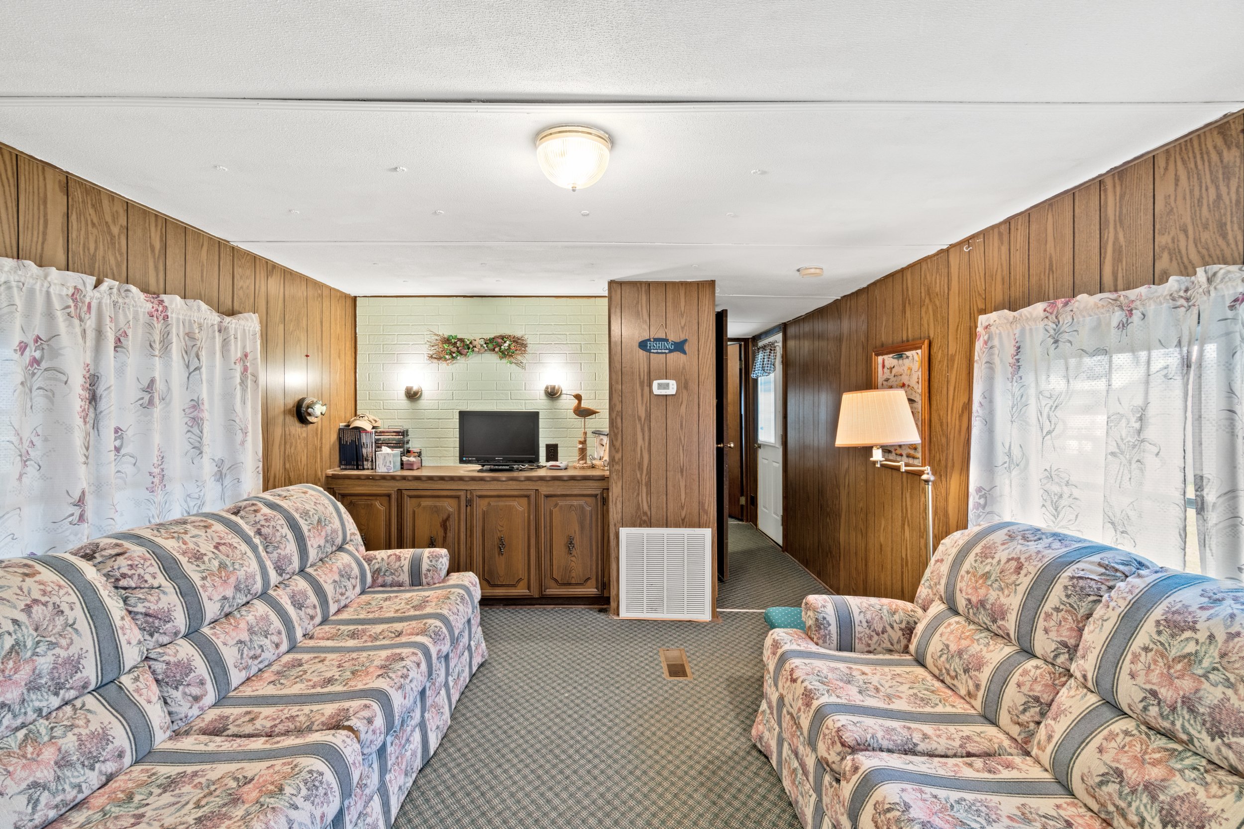 Living room with wood-paneled walls, floral couches, a small television on a wooden cabinet, and curtains with floral patterns, decorated with a lamp, picture, and fishing sign.