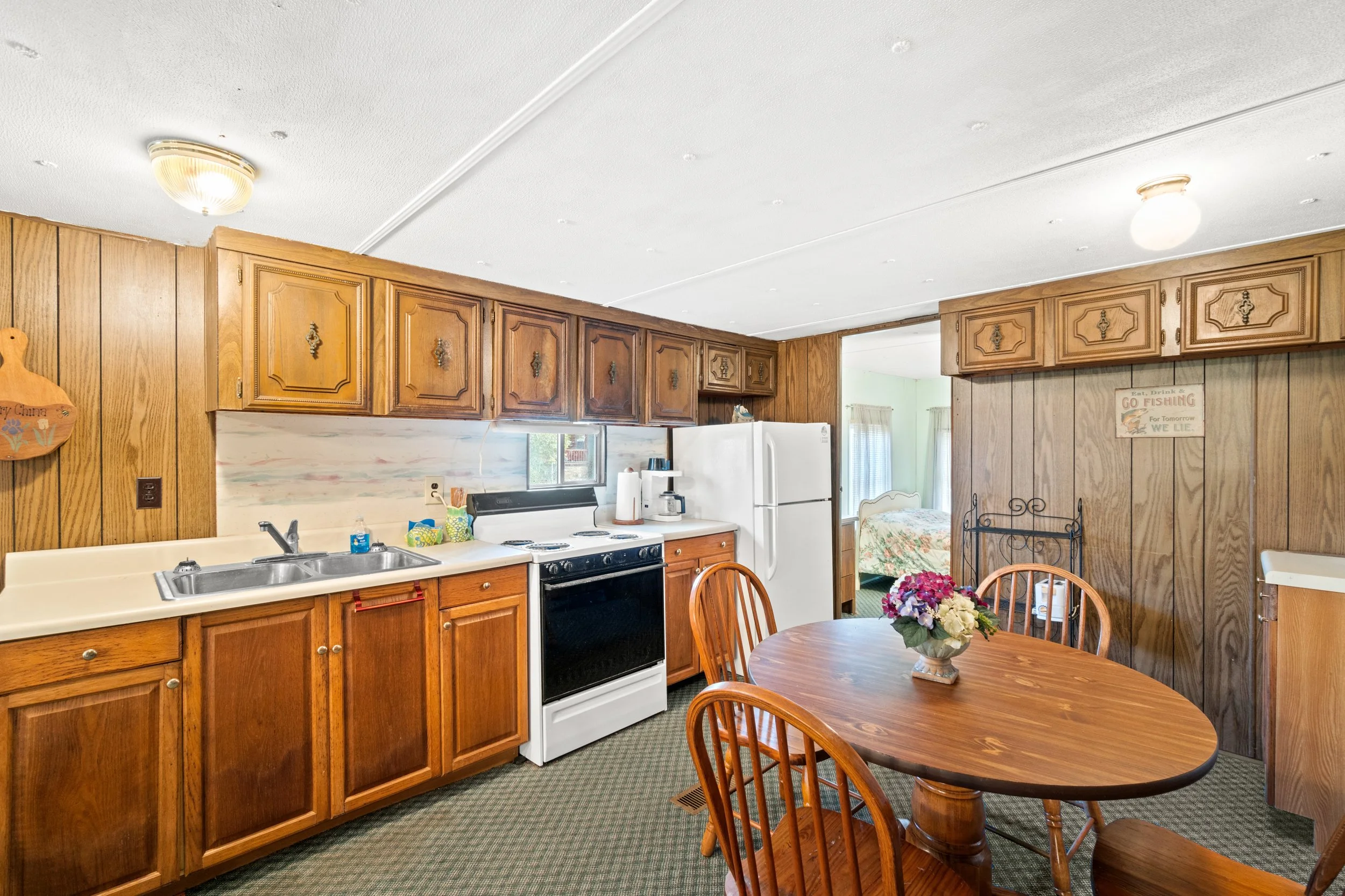Kitchen with wooden cabinets, white appliances, a round wooden dining table with four chairs, and a flower arrangement. A bedroom is visible through an open doorway.