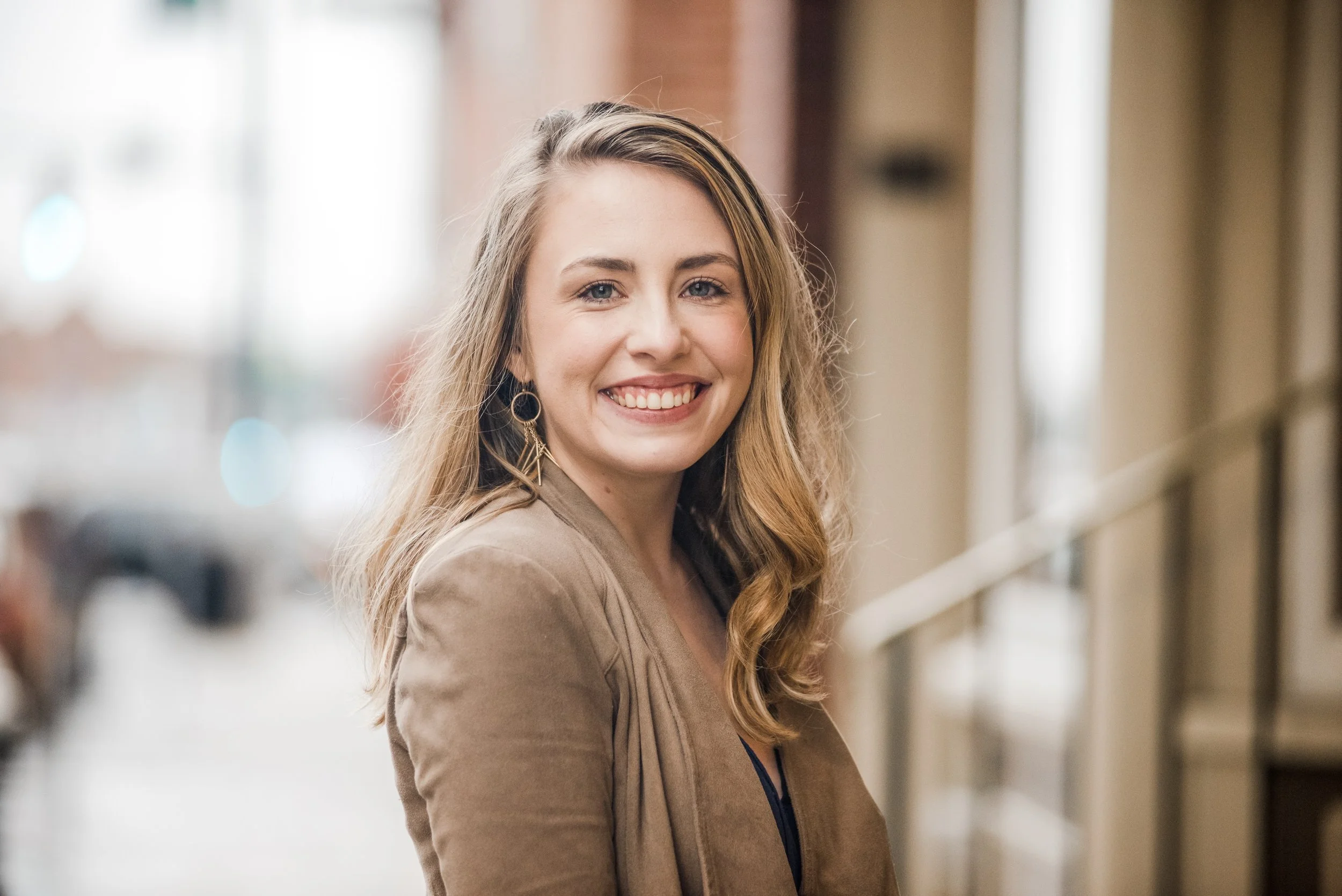 Nora Trotman smiling at the camera, wearing a tan blazer and earrings standing with a blurred city street background.