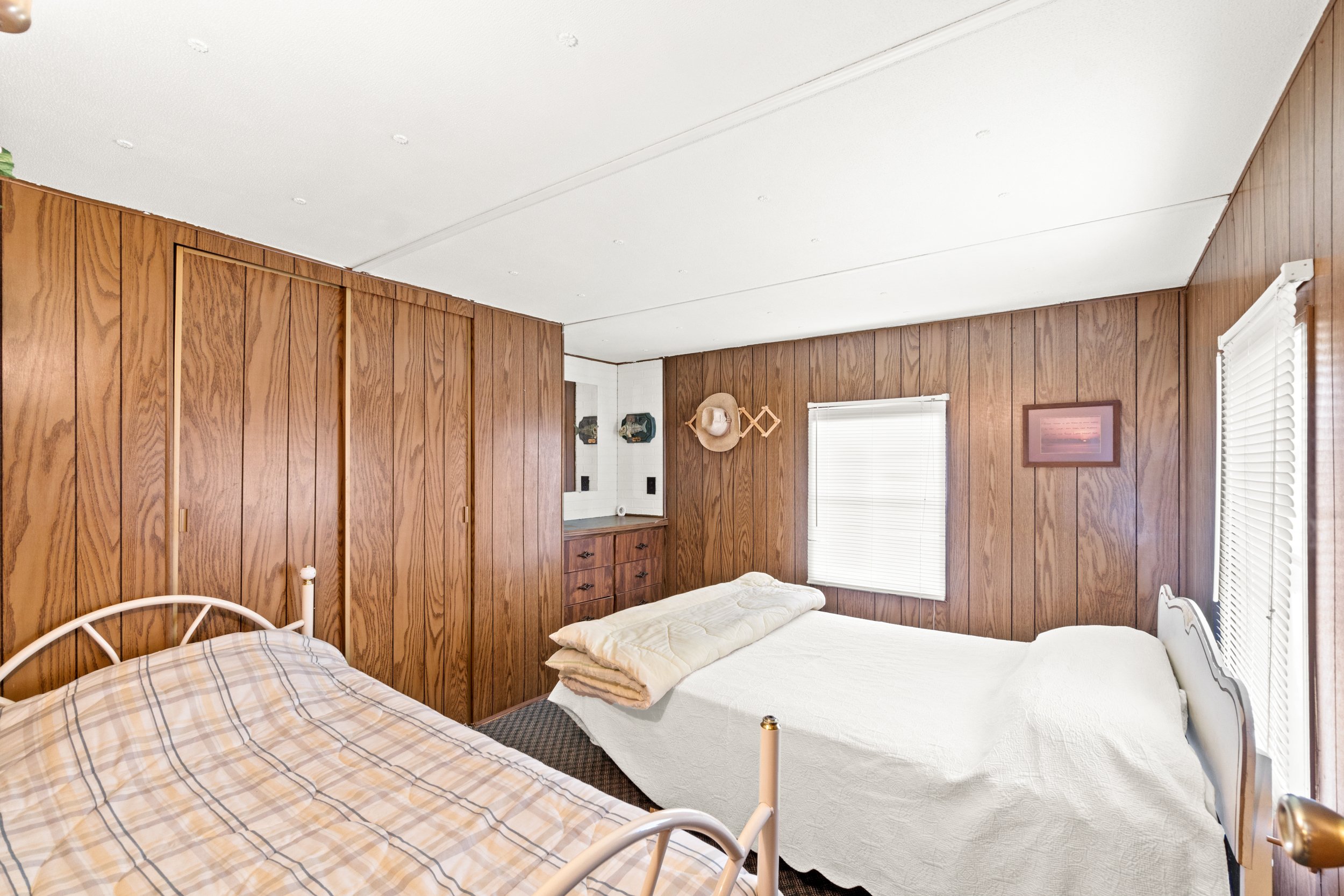 A bedroom with wood-paneled walls, two beds with white and beige bedding, a window with blinds, and wall decorations.
