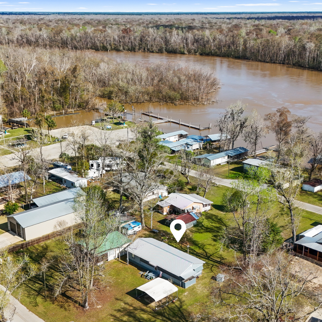Aerial view of a neighborhood near a large muddy river, with houses, trees, and docks along the shoreline. The area appears to be in early spring or late fall, with many trees without leaves.
