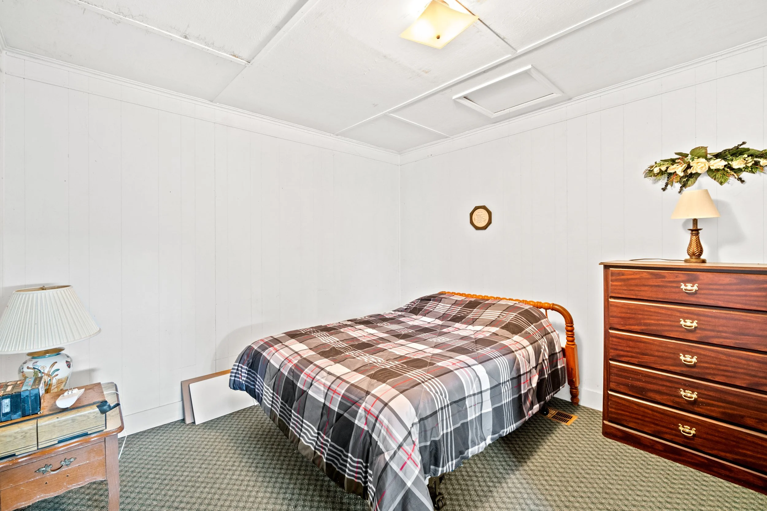A modest bedroom with white paneled walls, a bed with plaid bedding, a wooden dresser with a lamp and floral arrangement, a nightstand with a lamp and books, and a small framed picture on the wall.