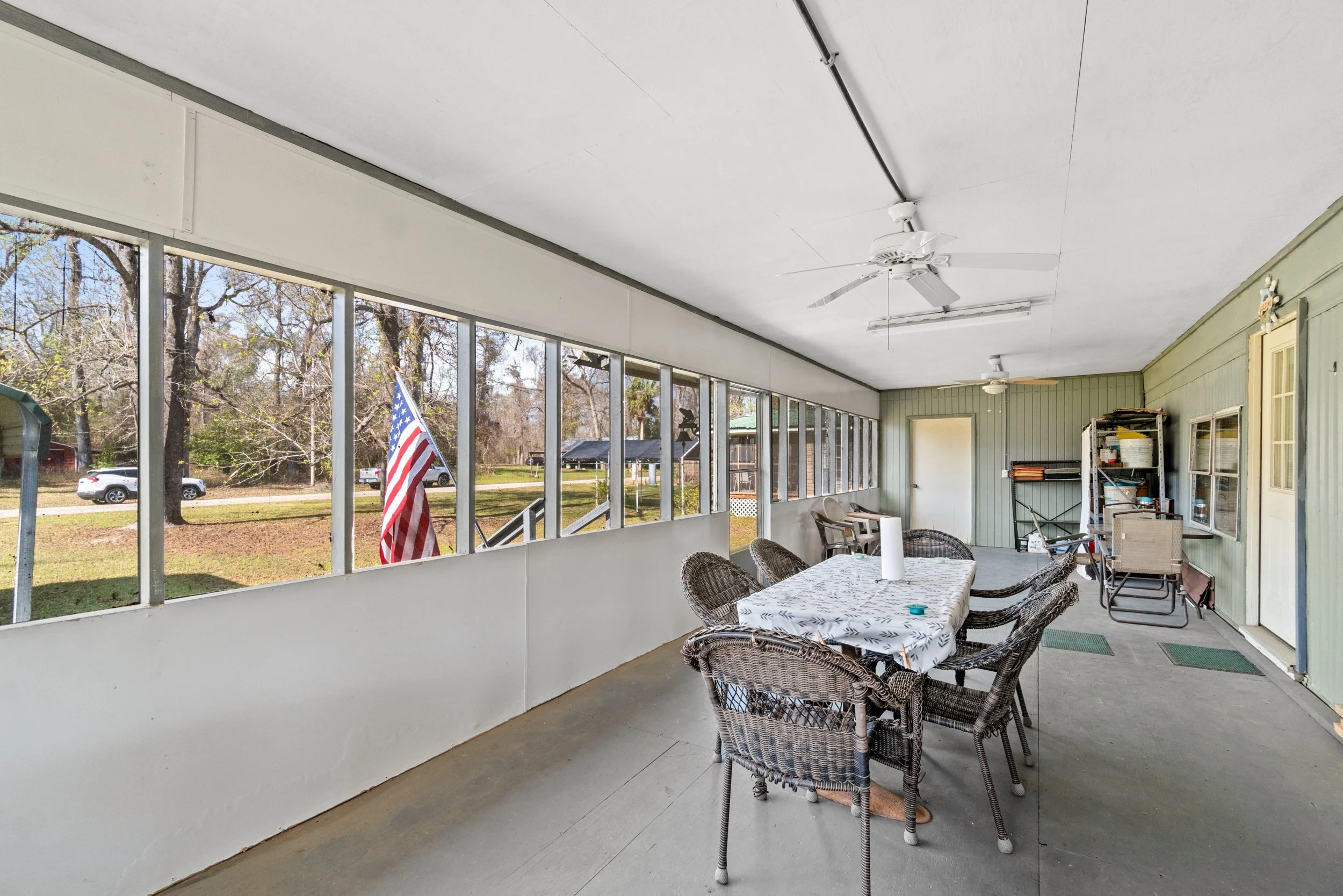 Enclosed porch with a dining table, wicker chairs, and shelving unit, overlooking trees and a yard outside