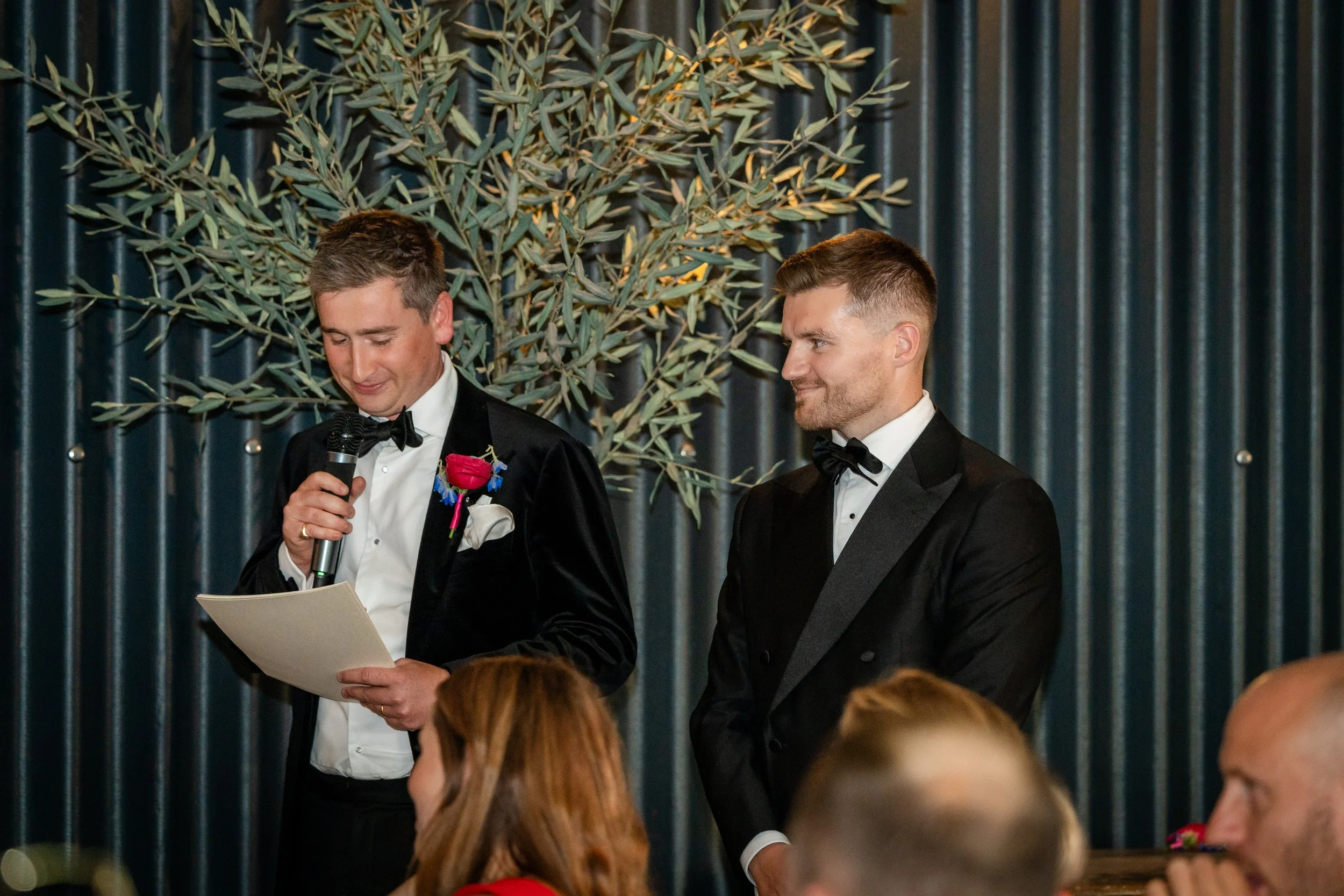 Two men in tuxedos standing at a formal event, one speaking into a microphone and holding a paper, the other looking on, with an audience and a backdrop of green plants and vertical wall panels.