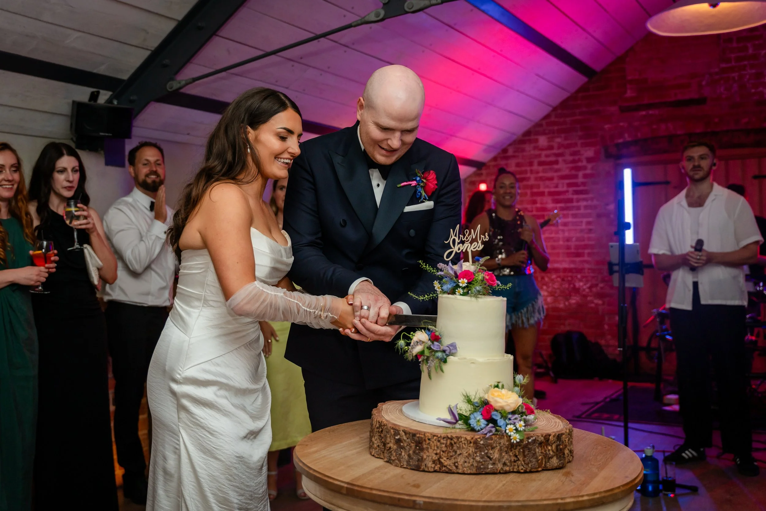 Bride and groom cutting a wedding cake together at a celebration, with guests watching and smiling around them.