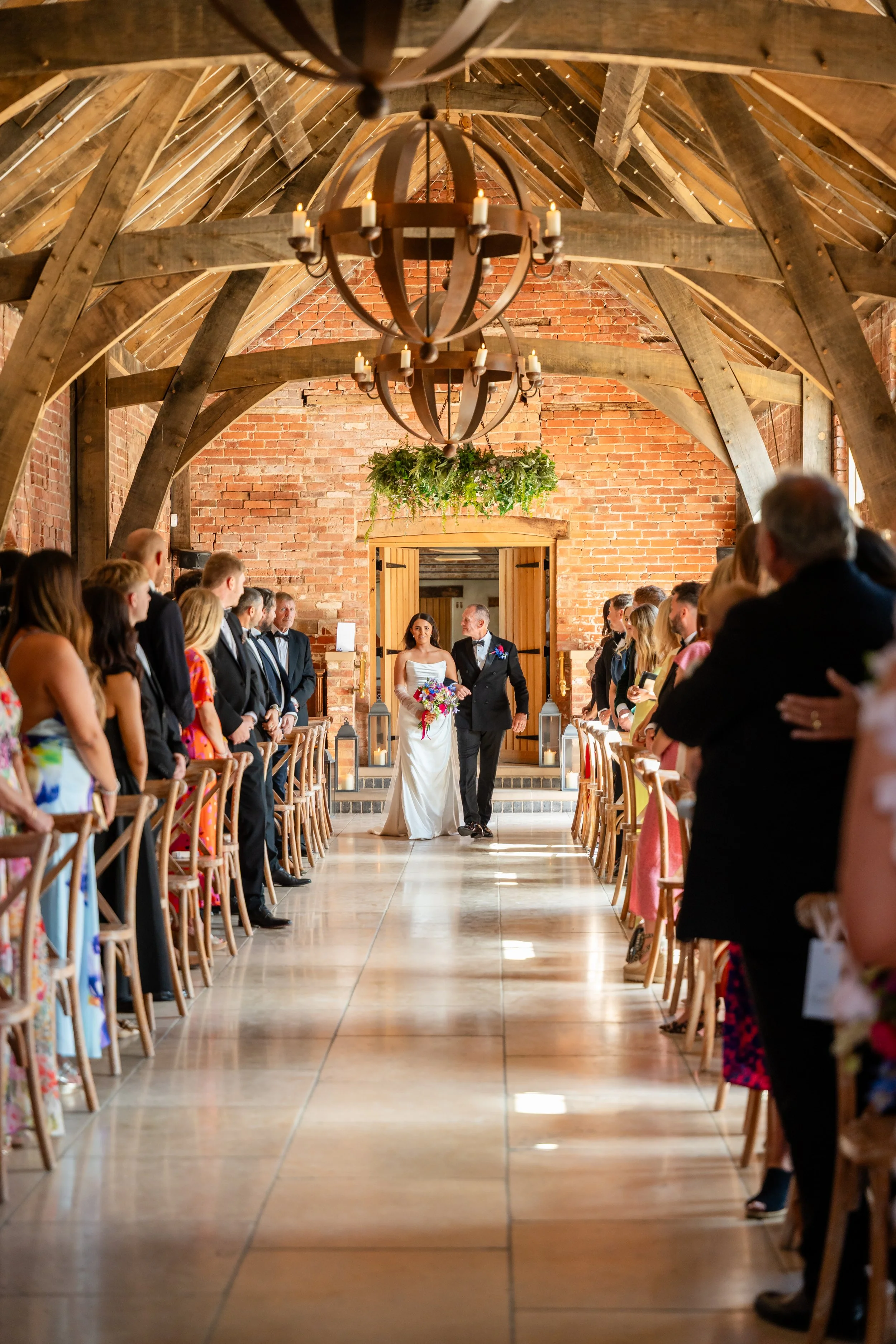 Wedding ceremony inside a rustic barn with wooden beams, brick walls, chandeliers, and guests standing on either side of the aisle as a bride and groom walk down the aisle.