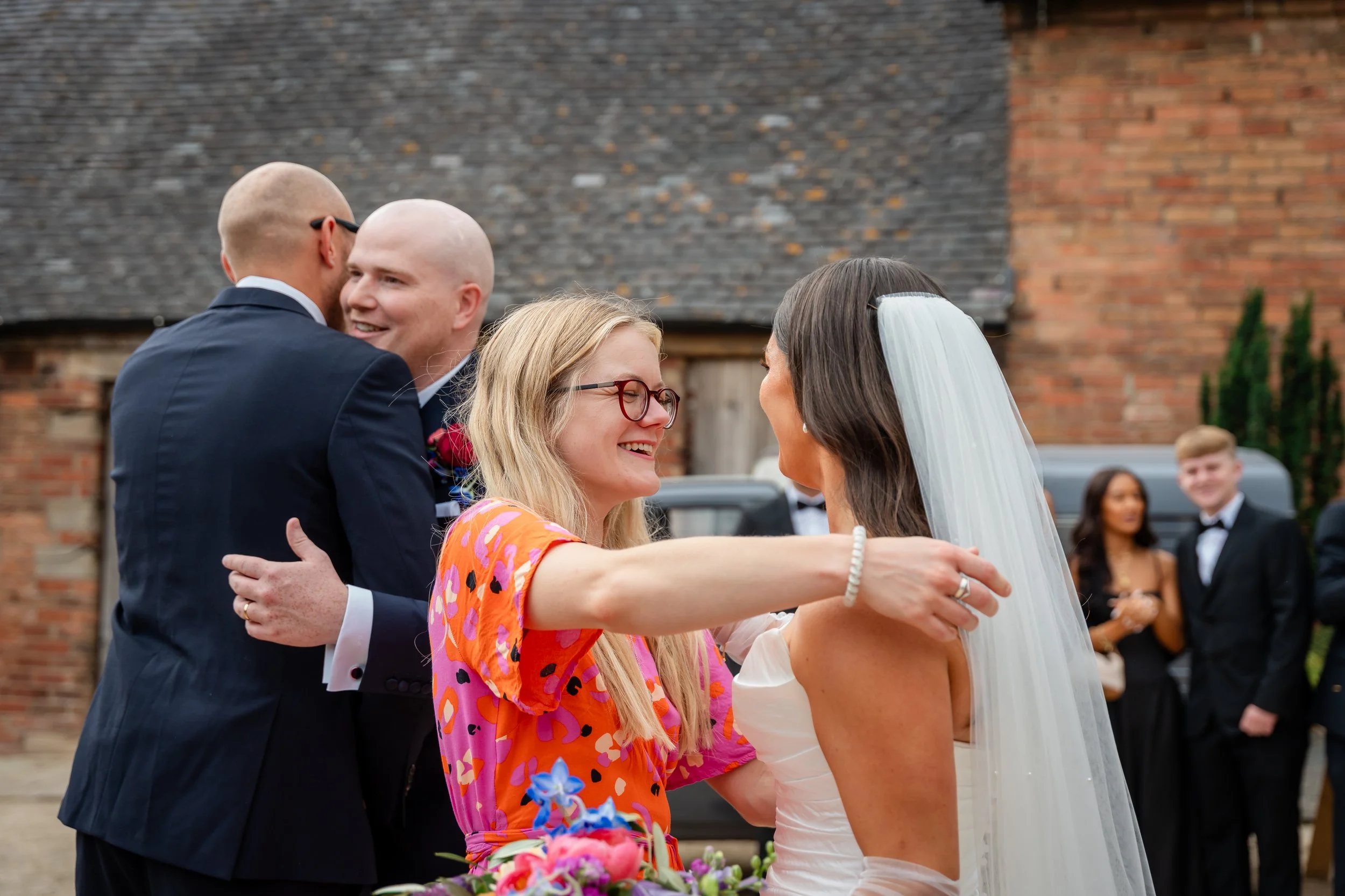People hugging at a wedding reception outdoors, with a brick building in the background.