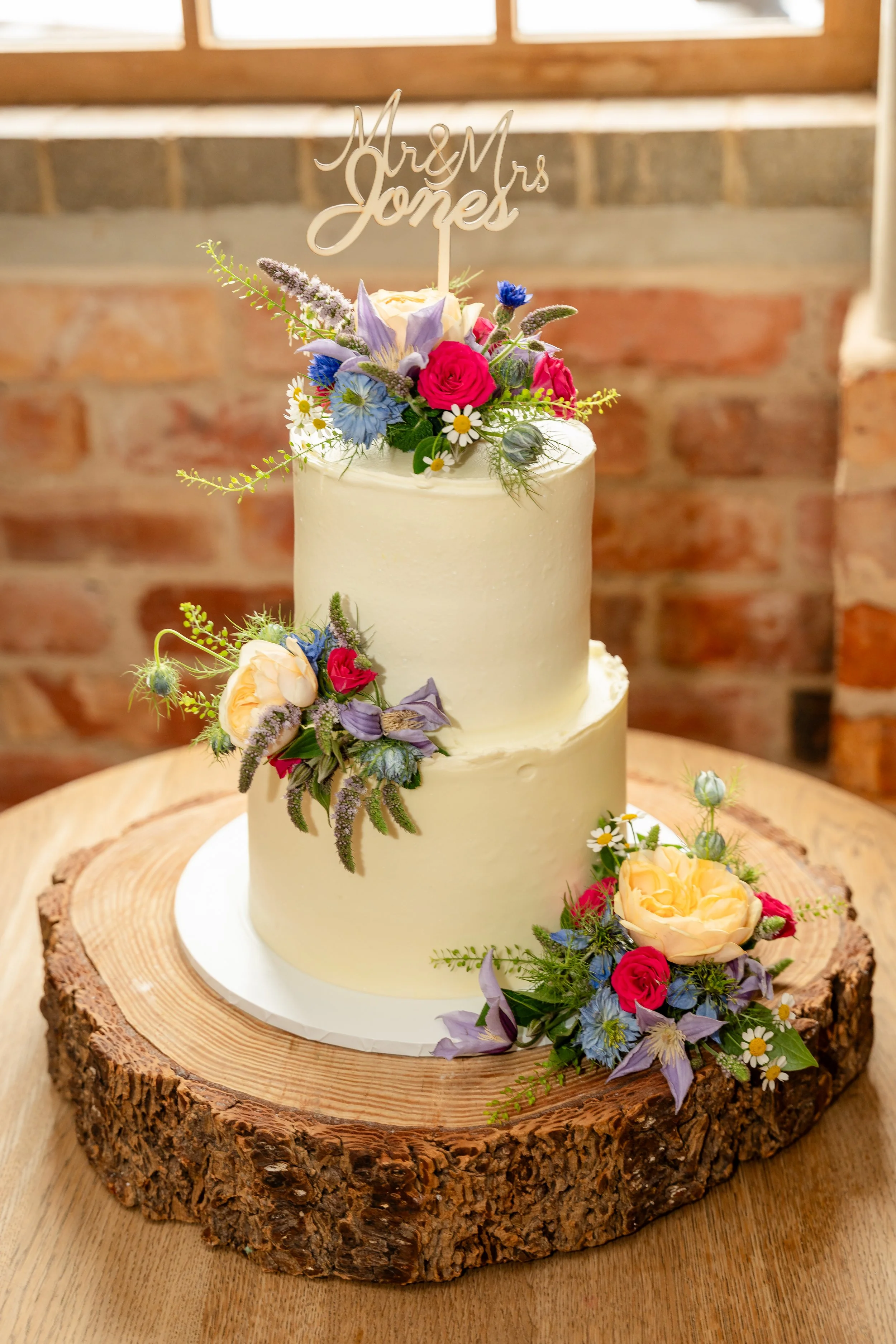 A wedding cake decorated with colorful fresh flowers and a cake topper that reads 'Mr & Mrs Jones', placed on a rustic wooden slab.