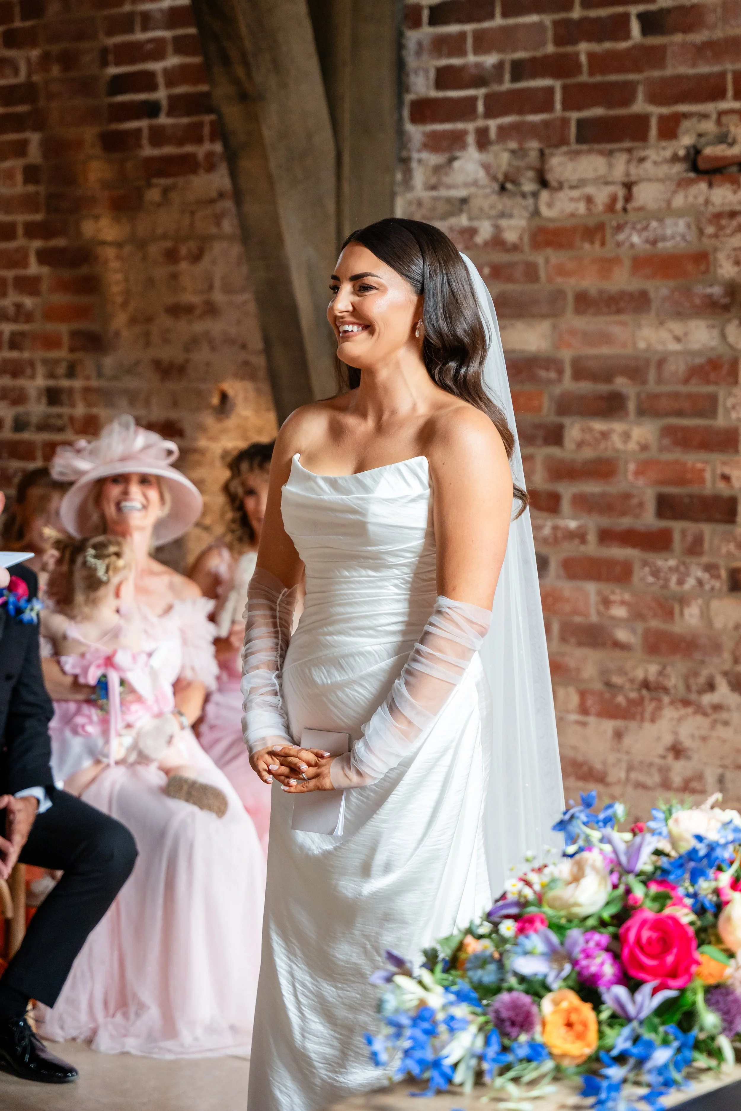 A bride in a white wedding gown and veil stands smiling in front of a brick wall during a wedding ceremony, with guests and colorful flowers visible in the background.