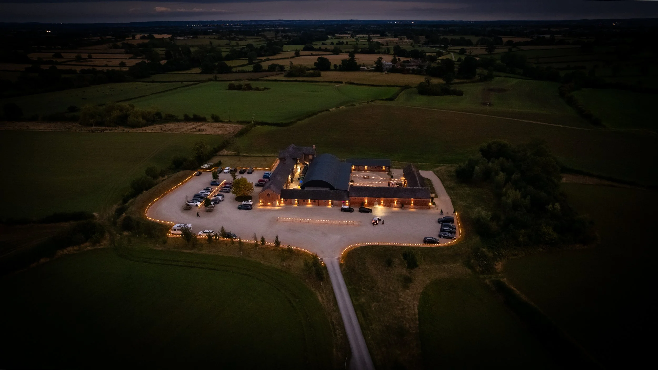 An aerial night view of a rural estate with a lit building, surrounded by a parking lot with cars, and lush green fields extending into the distance.