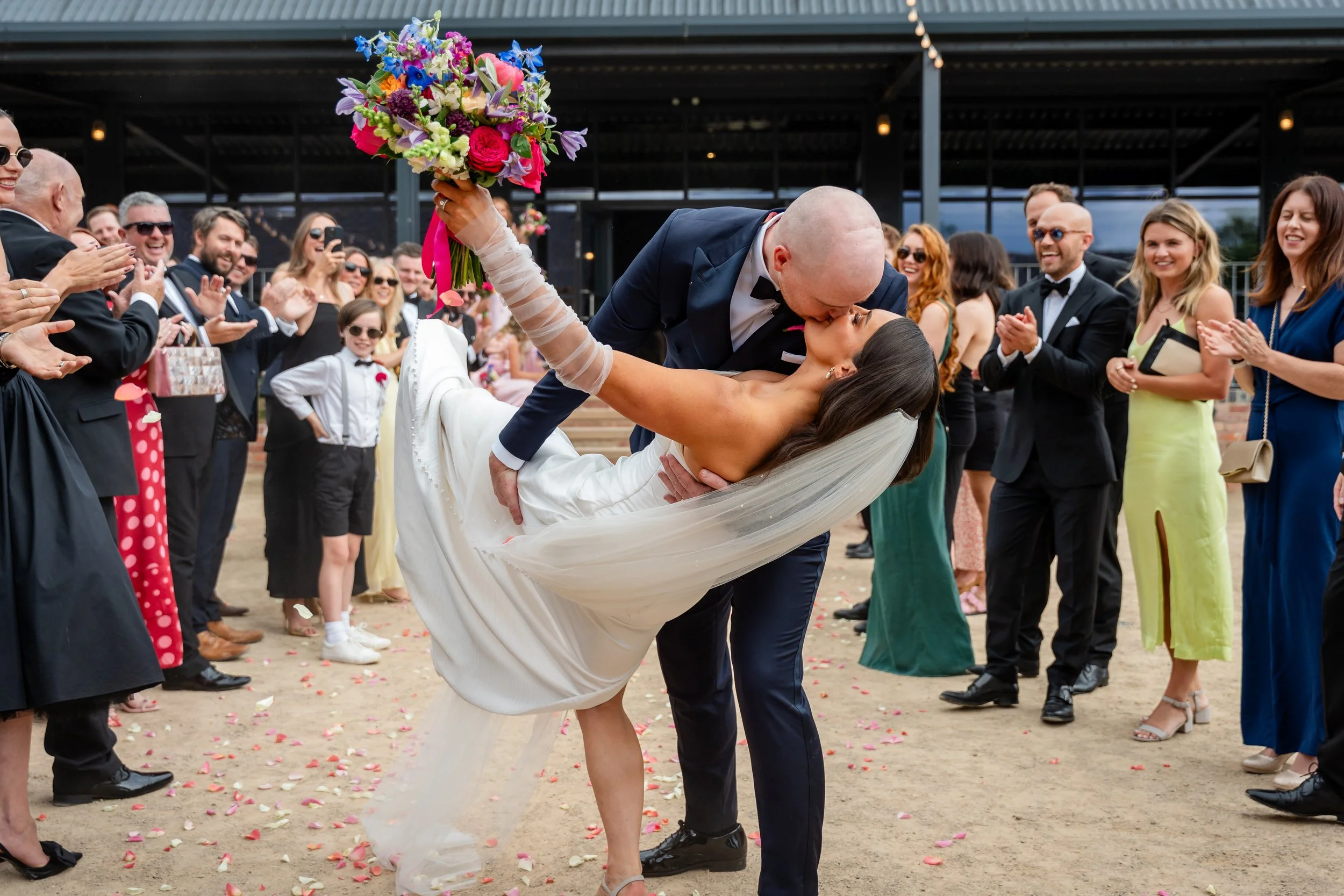 A newlywed couple sharing a kiss at their wedding reception. The groom, dressed in a navy suit, is leaning over the bride, who is in a white wedding dress, as he kisses her. The bride is holding a vibrant bouquet of flowers. Guests in formal attire s