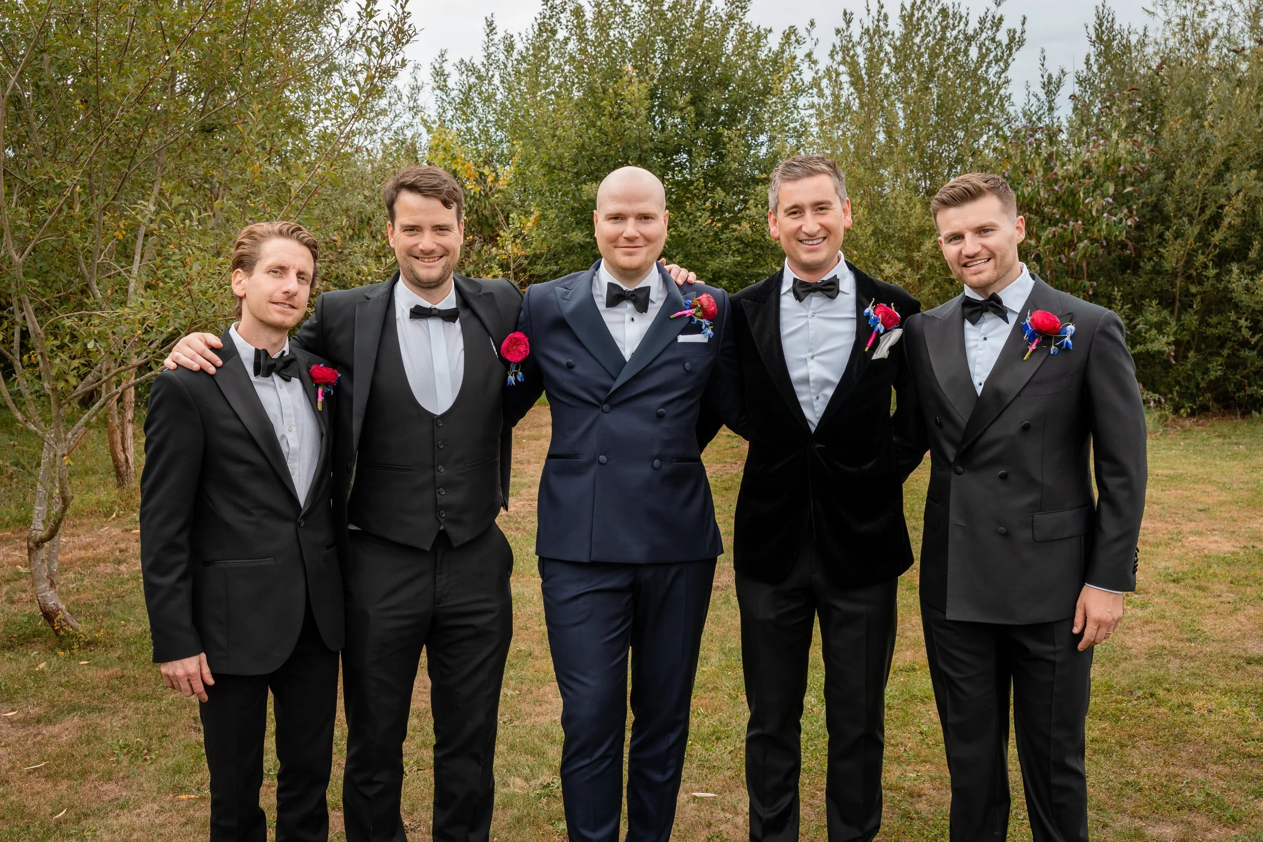 Group of five men in tuxedos standing outdoors among trees, smiling at the camera, with one man in the middle wearing a navy blue tuxedo, at an outdoor event.