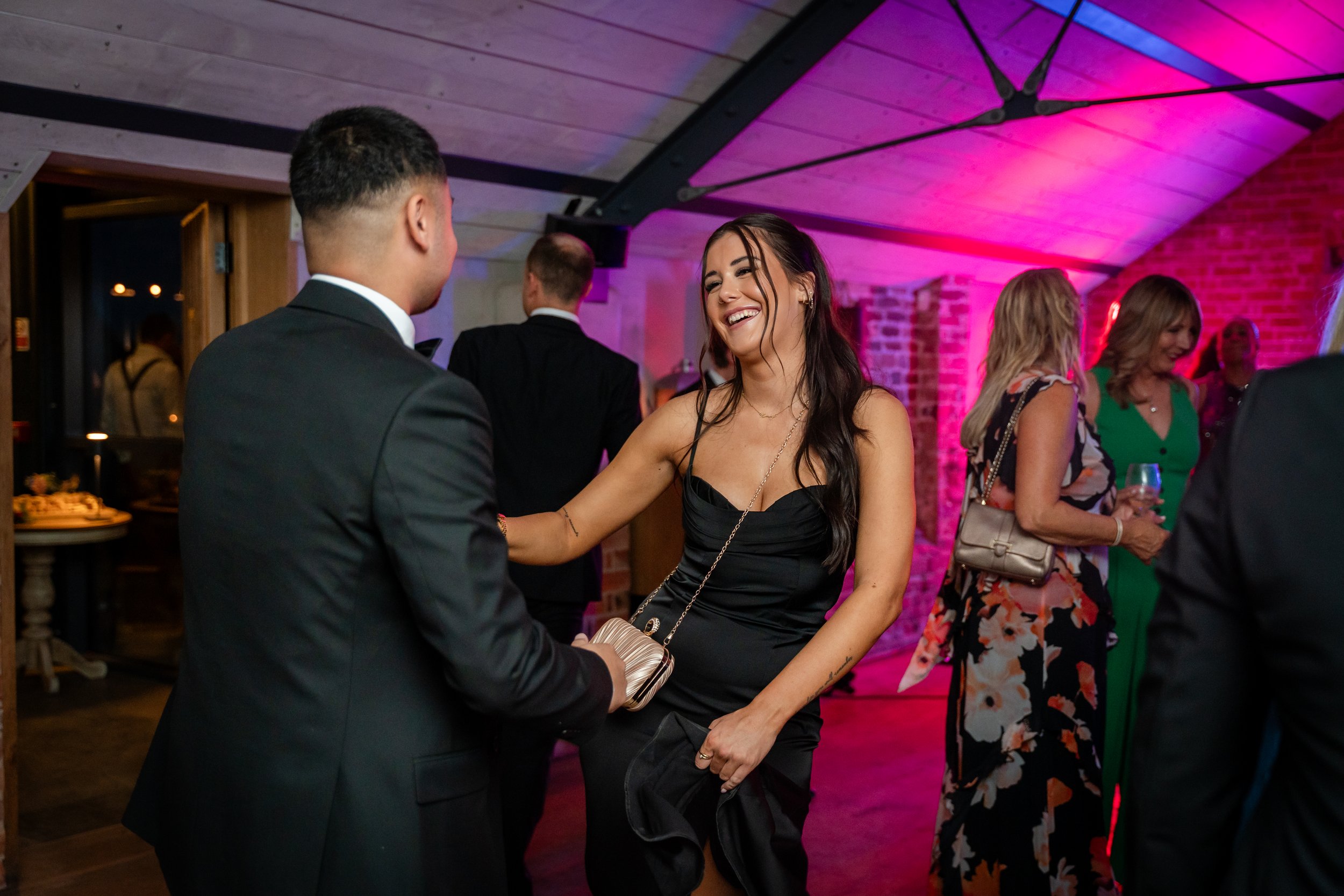 A woman and a man dancing at a social event, with other people in the background, under pink and purple lighting, in a venue with brick walls and a wooden ceiling.