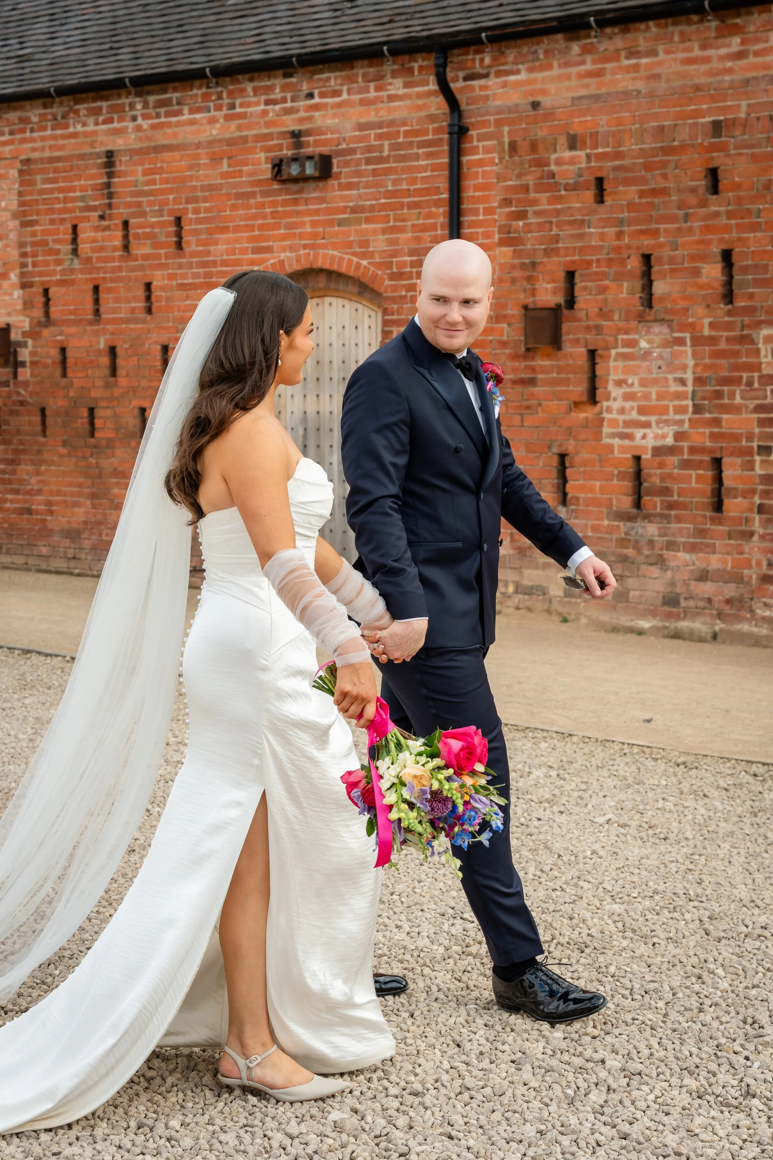 A newlywed couple walking outside holding hands, with a woman in a white wedding dress and veil and a man in a navy suit holding a colorful bouquet of flowers, against a red brick wall.
