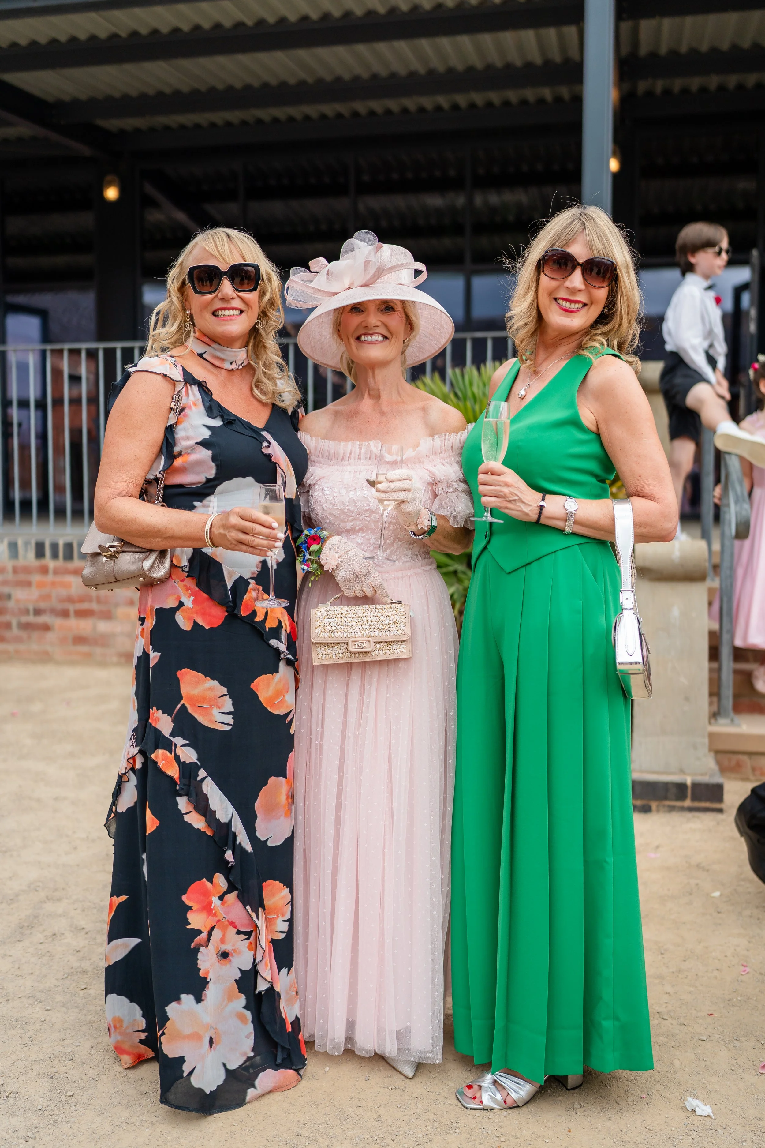 Three women dressed elegantly, smiling and holding glasses of champagne at a social event, with a young boy in the background.