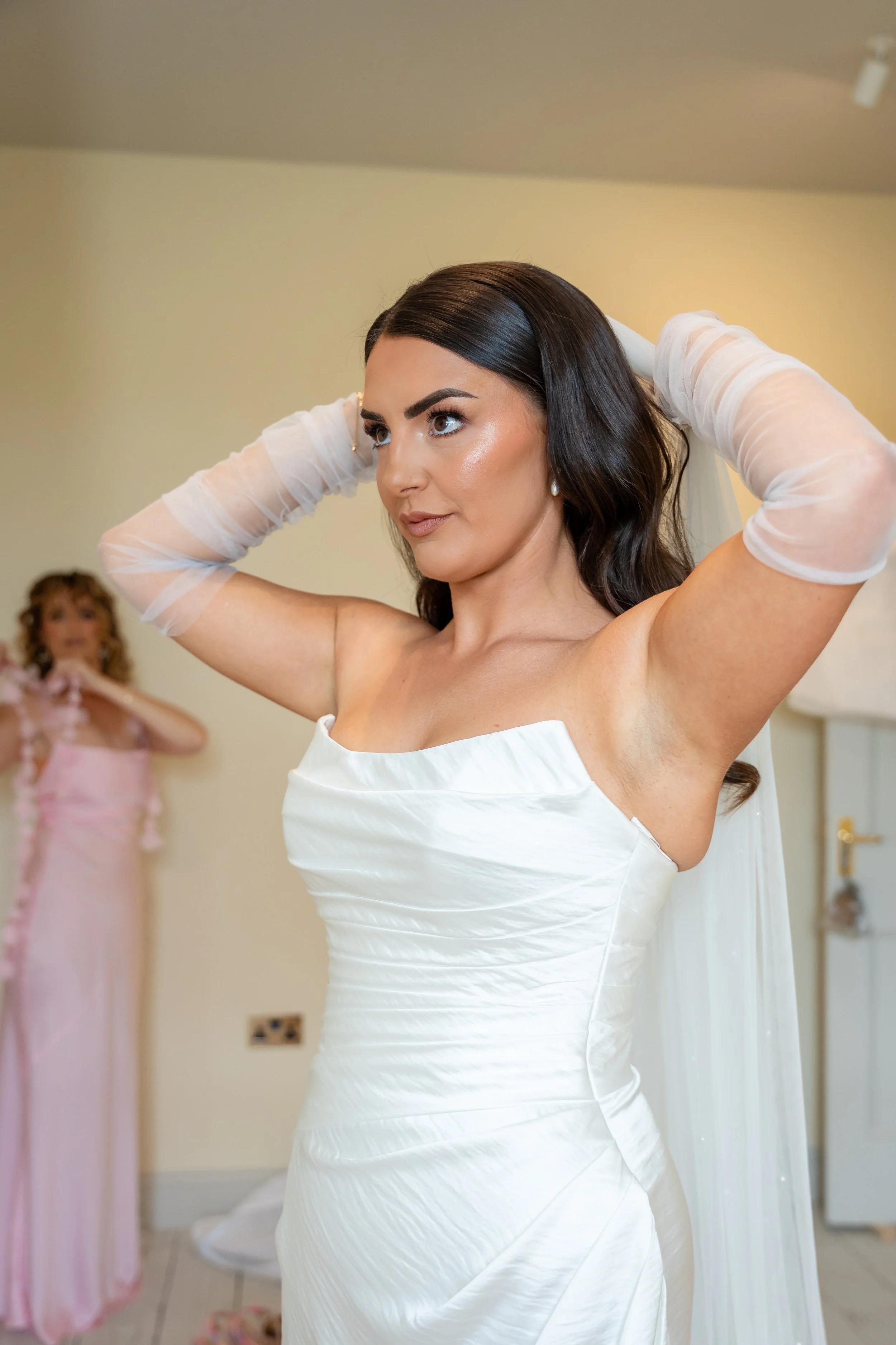 A woman in a white dress is adjusting her hair in a room, with another woman in a pink dress blurred in the background.