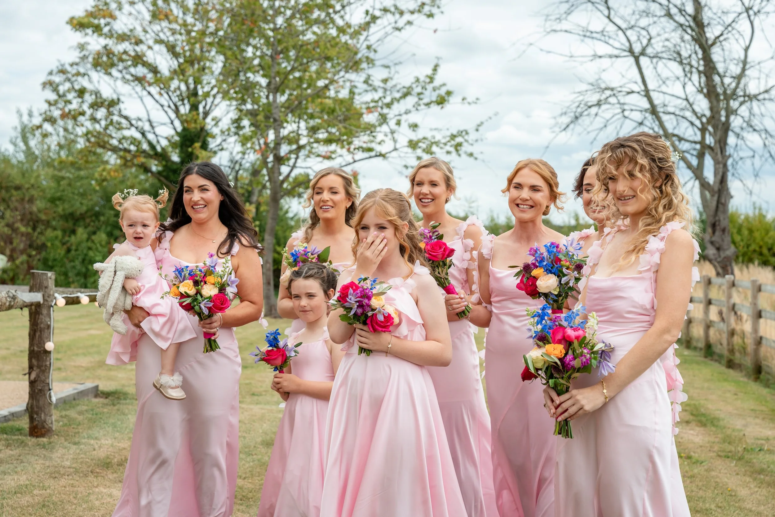 Group of women and girls dressed in pink, holding colorful bouquets, some smiling and some surprised, outdoors during a cloudy day.