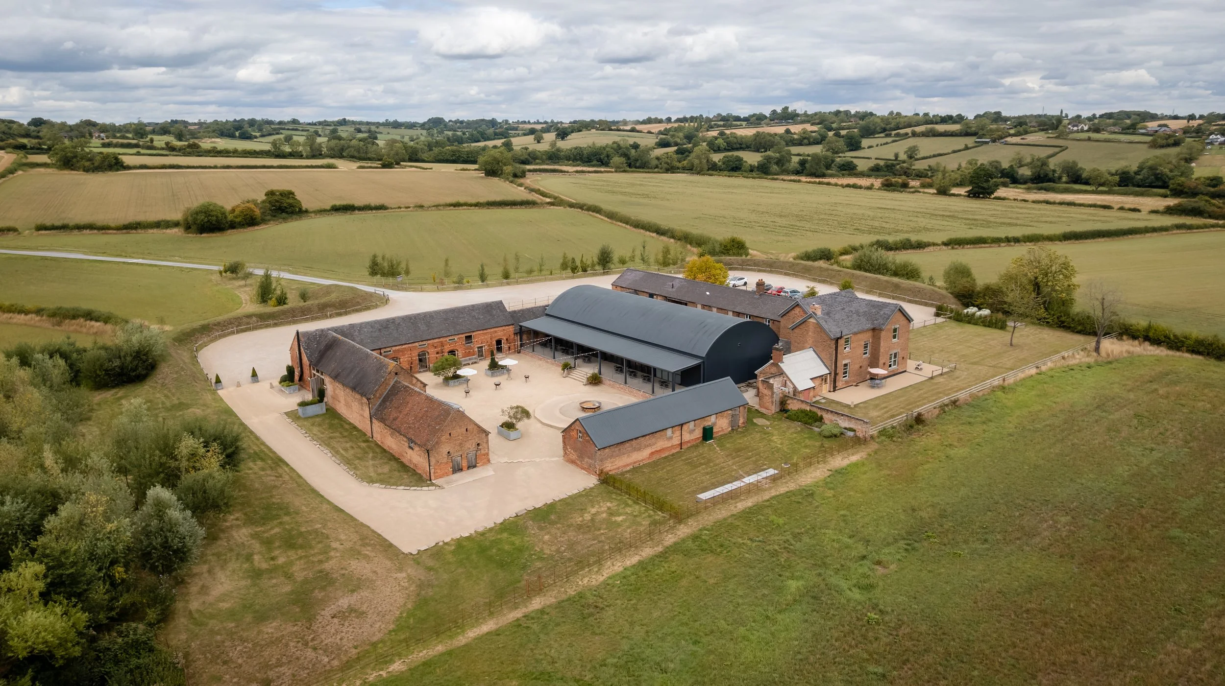 Aerial view of a rural estate with brick buildings, some with metal roofs, surrounded by green fields and trees, under a cloudy sky.