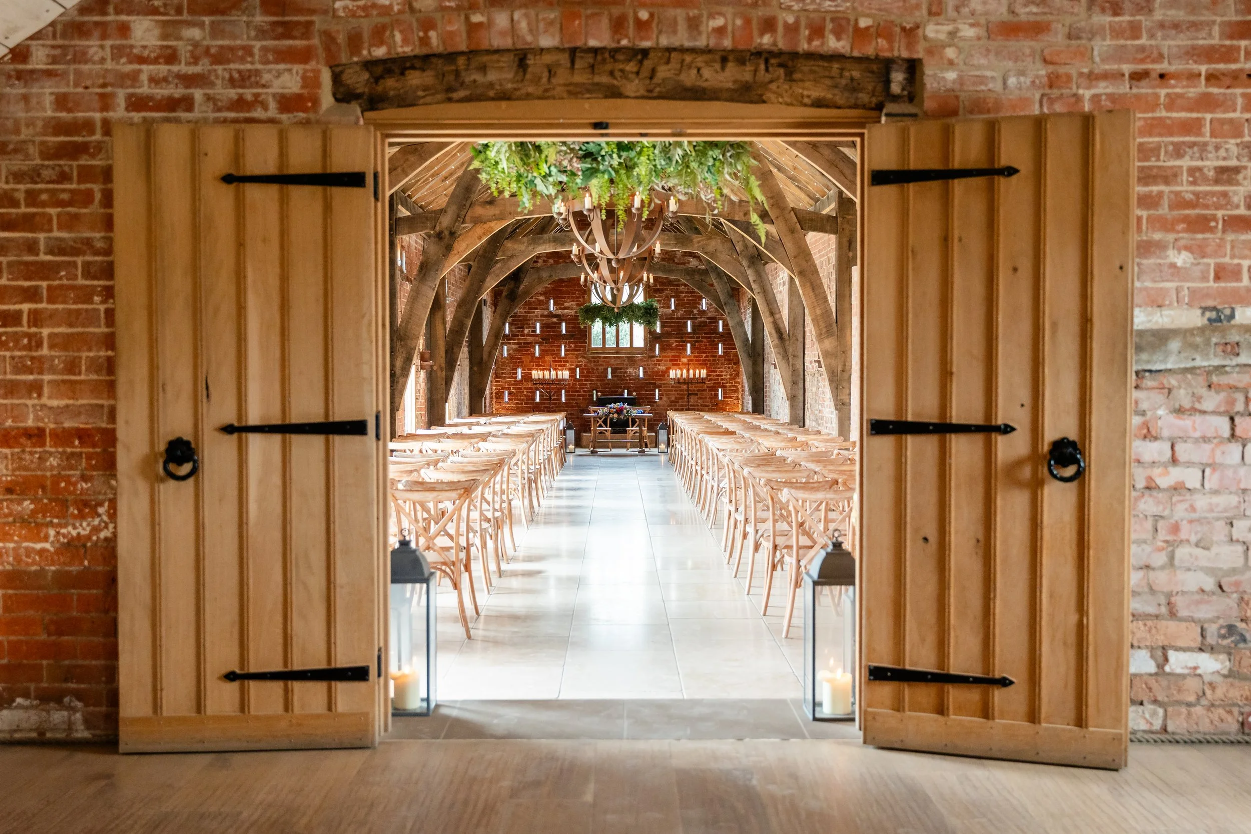 View of an indoor wedding or event space through wooden door with black hinges, with brick walls, rustic wooden beams, chandeliers, greenery, and rows of wooden chairs arranged facing an altar with candles.