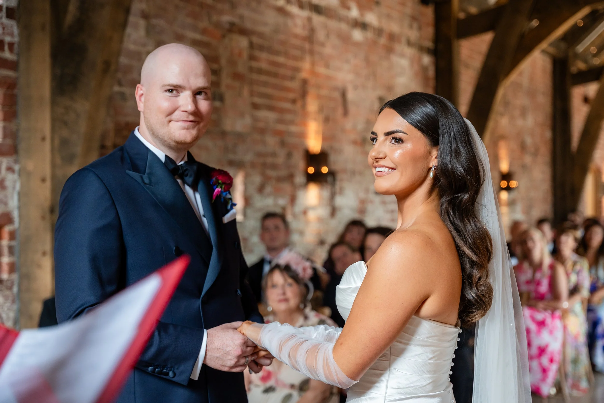 A bride and groom exchanging vows during a wedding ceremony in an indoor venue with exposed brick walls and wooden beams.