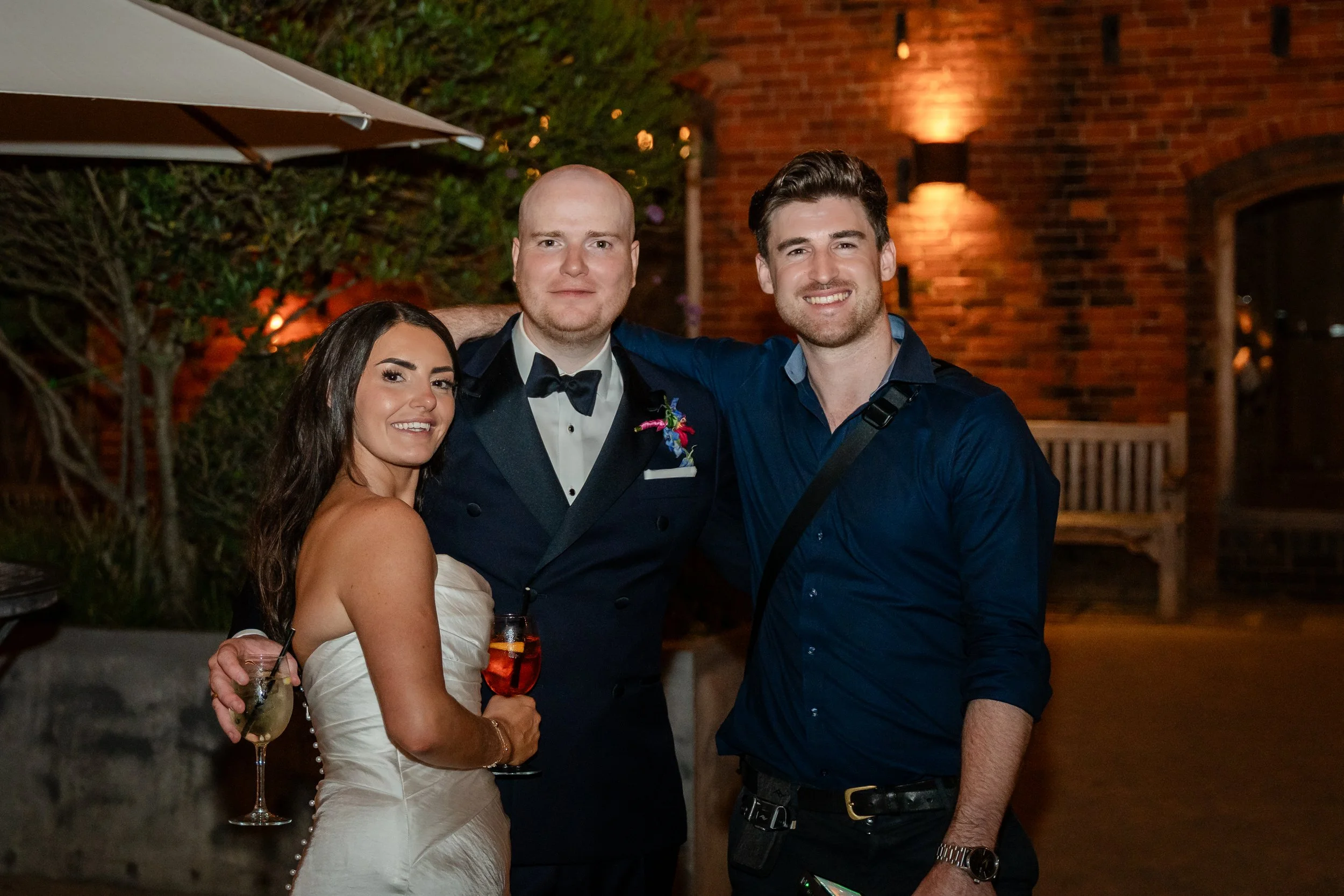 Group of four friends smiling at a nighttime outdoor event, standing close together, with drinks in hand, in front of a brick building with warm lighting.