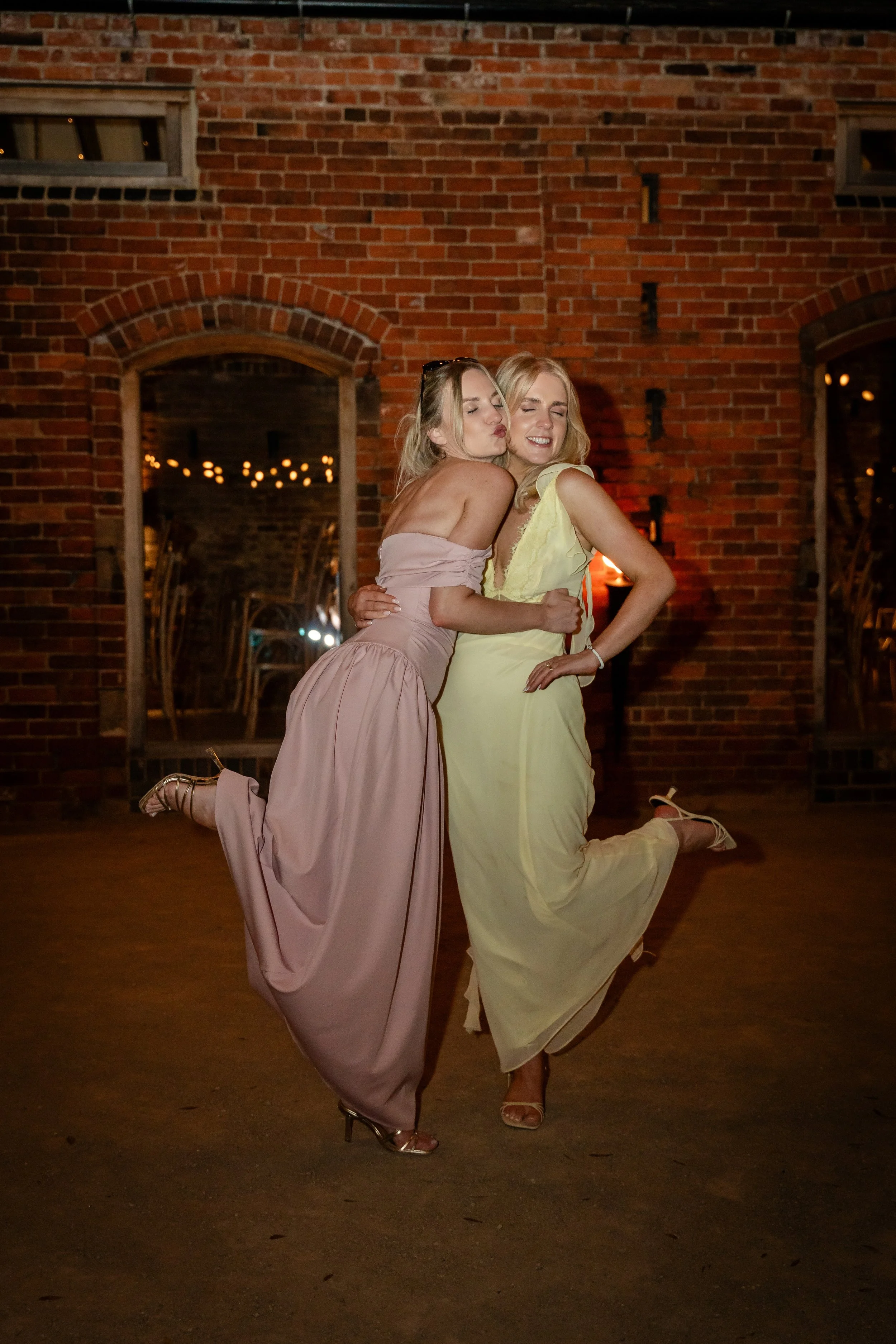 Two women in formal dresses posing playfully indoors with a brick wall background at night.