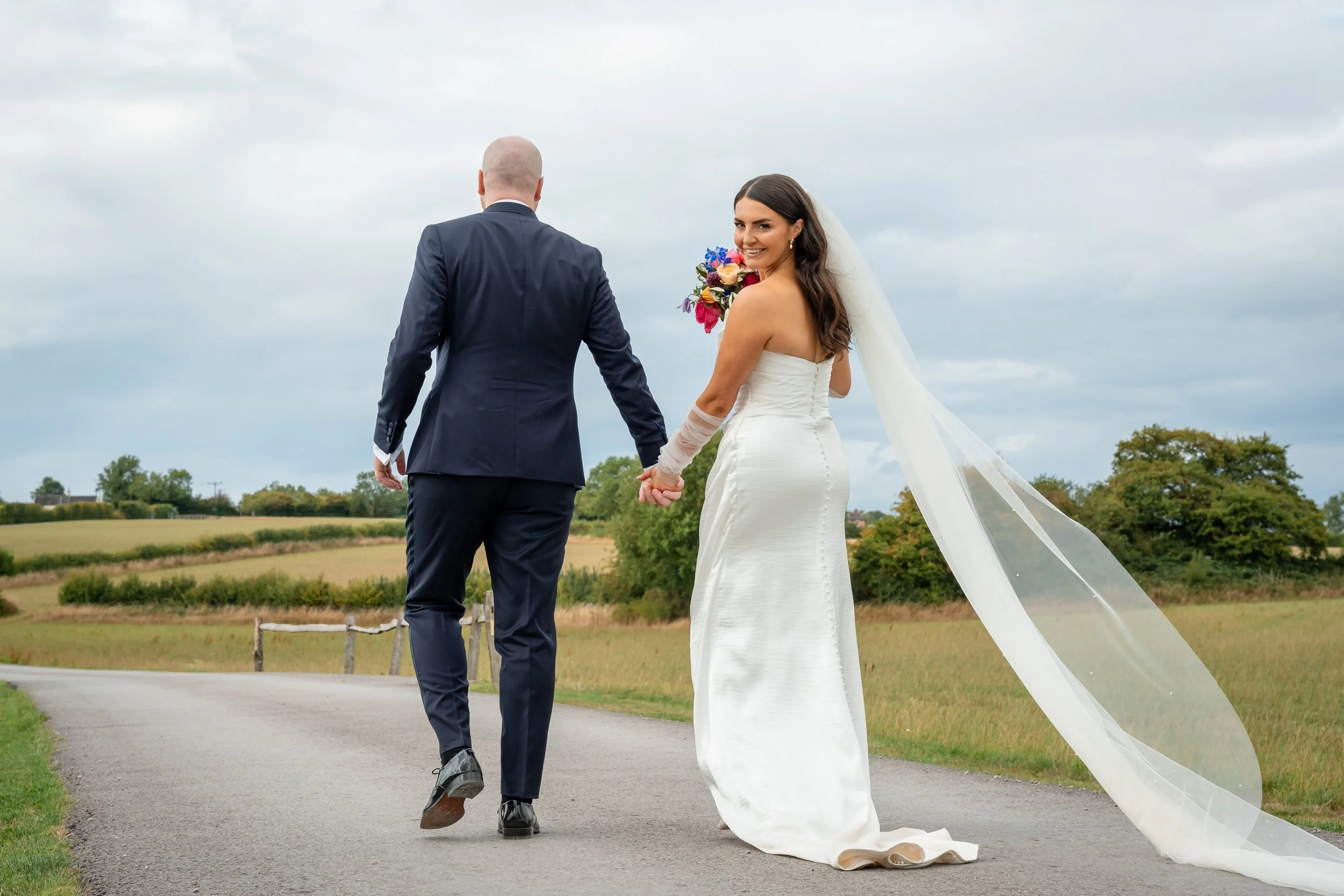 A bride in a white strapless wedding gown with a long veil holding hands with a groom in a dark suit walking on a rural road with green fields and trees around, under a cloudy sky.