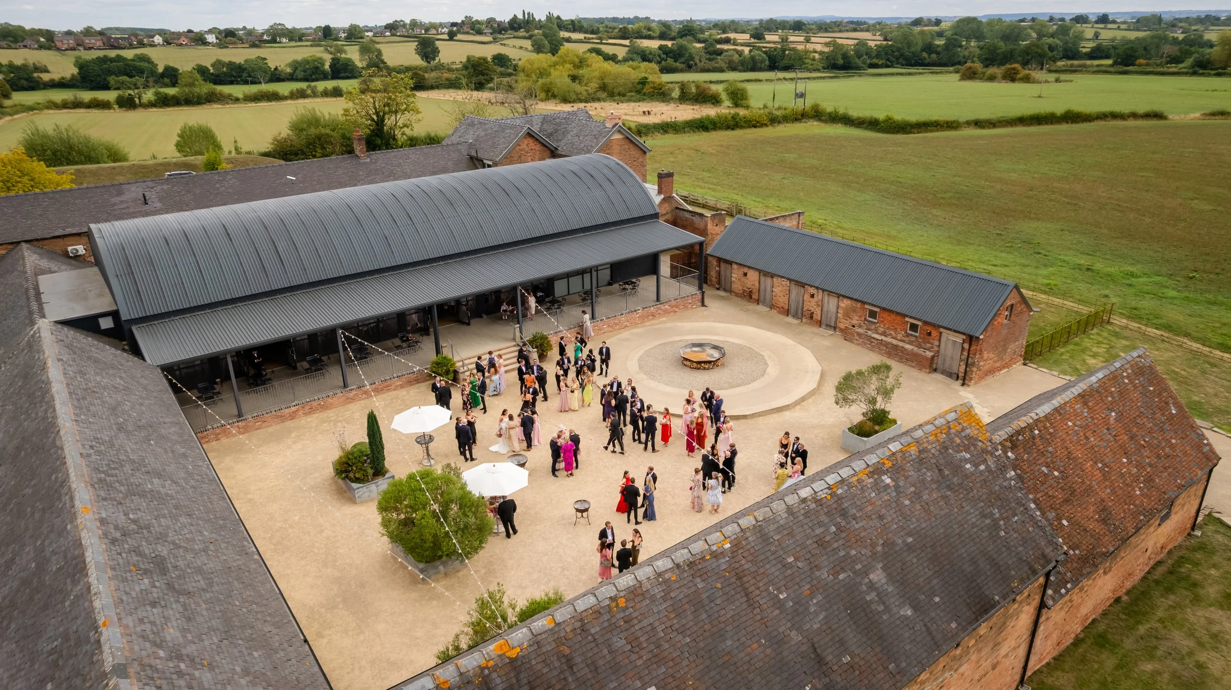 An outdoor wedding reception with guests gathered around a circular fire pit at a rustic barn venue, surrounded by green fields and rolling countryside.