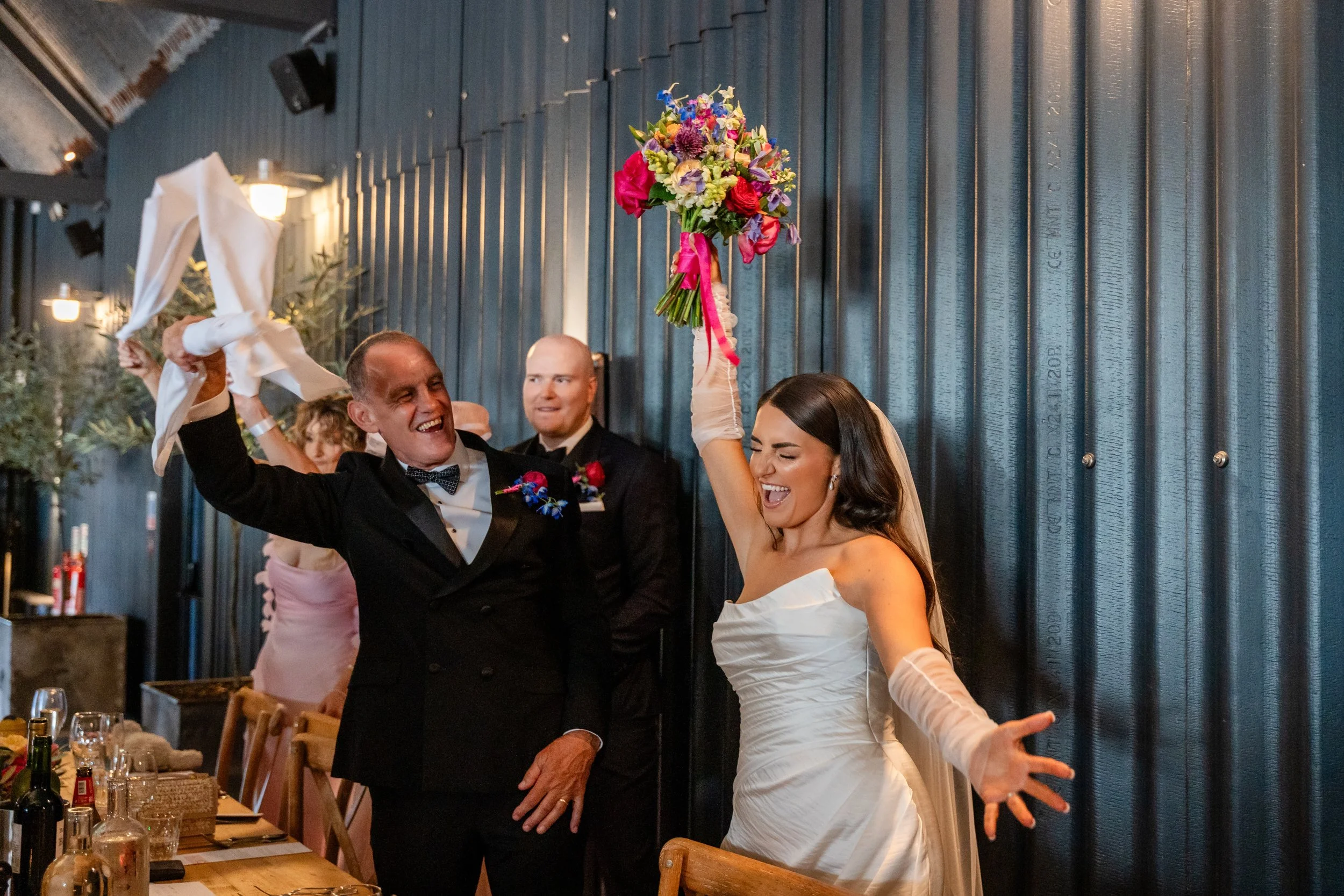 A joyful man in a tuxedo and a woman in a white wedding dress celebrating at a wedding reception. The woman is holding a colorful bouquet of flowers high above her head. They are dancing and smiling.